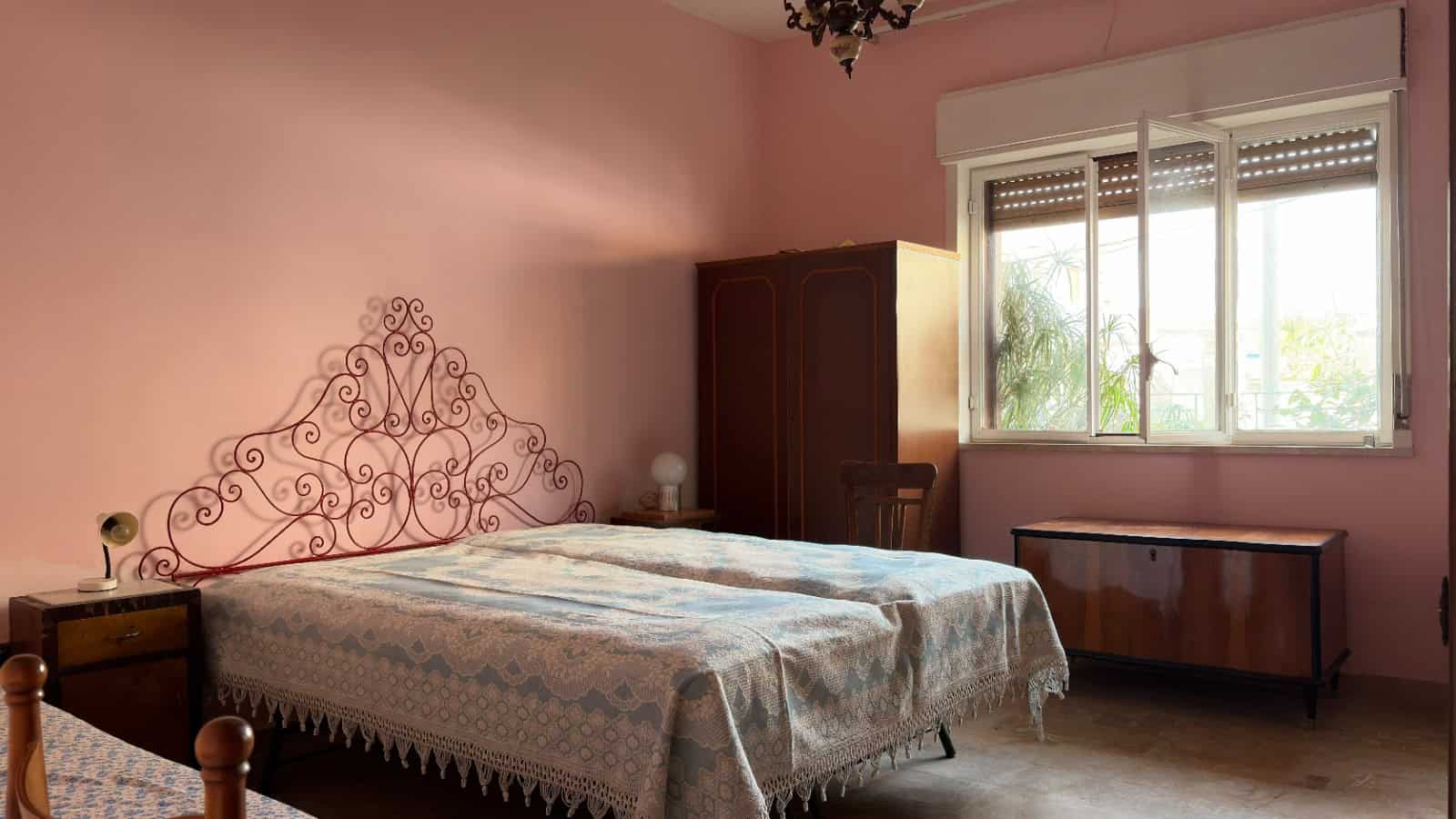 Ampia camera da letto con pareti rosa, letto matrimoniale con testiera in ferro battuto, armadio in legno e finestra che lascia entrare luce naturale, ideale per case e appartamenti in vendita o affit.