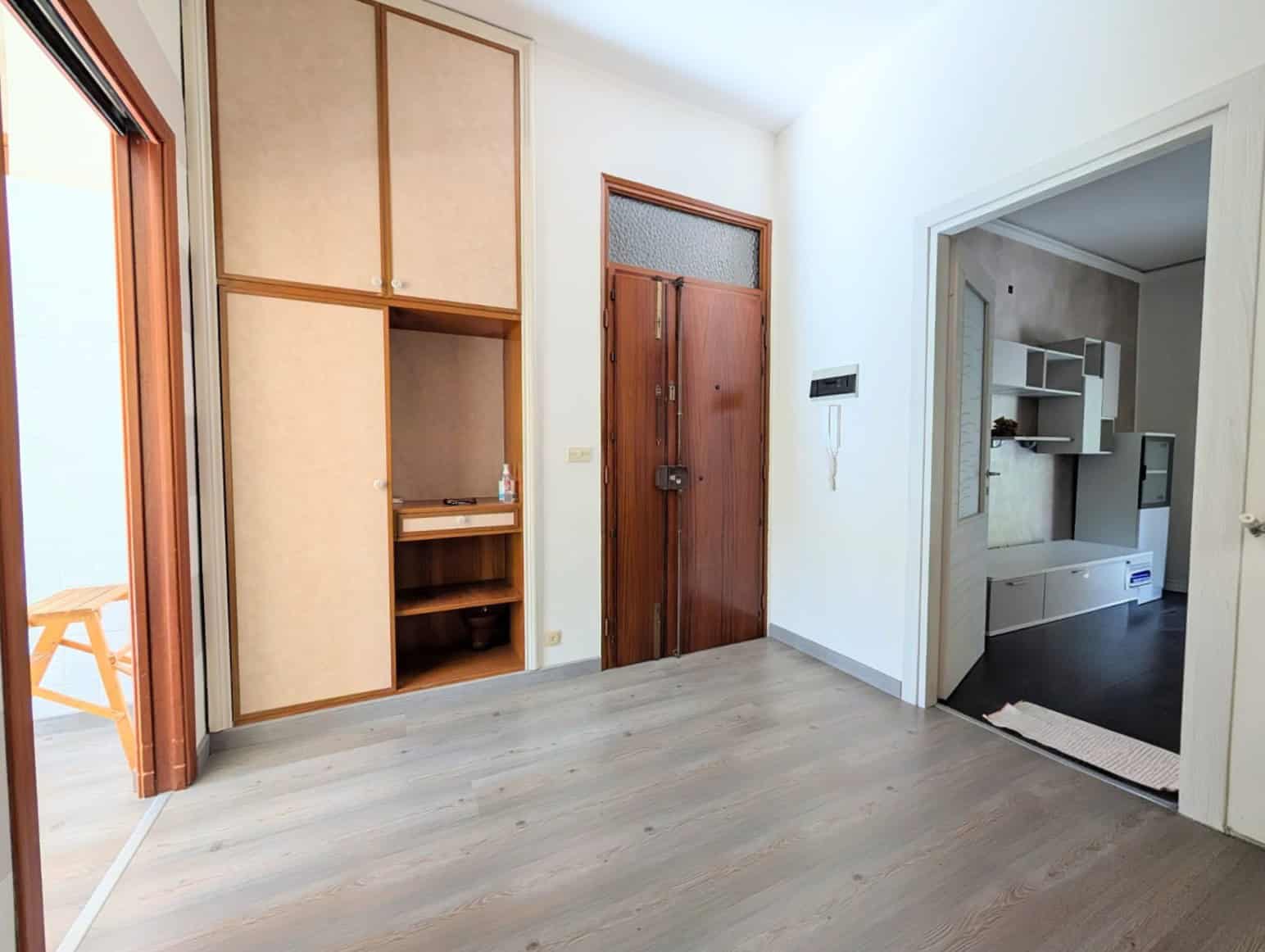 Interno di un appartamento con pareti bianche, porta in legno e spazio di archiviazione, con accesso a una cucina moderna e luminosa.