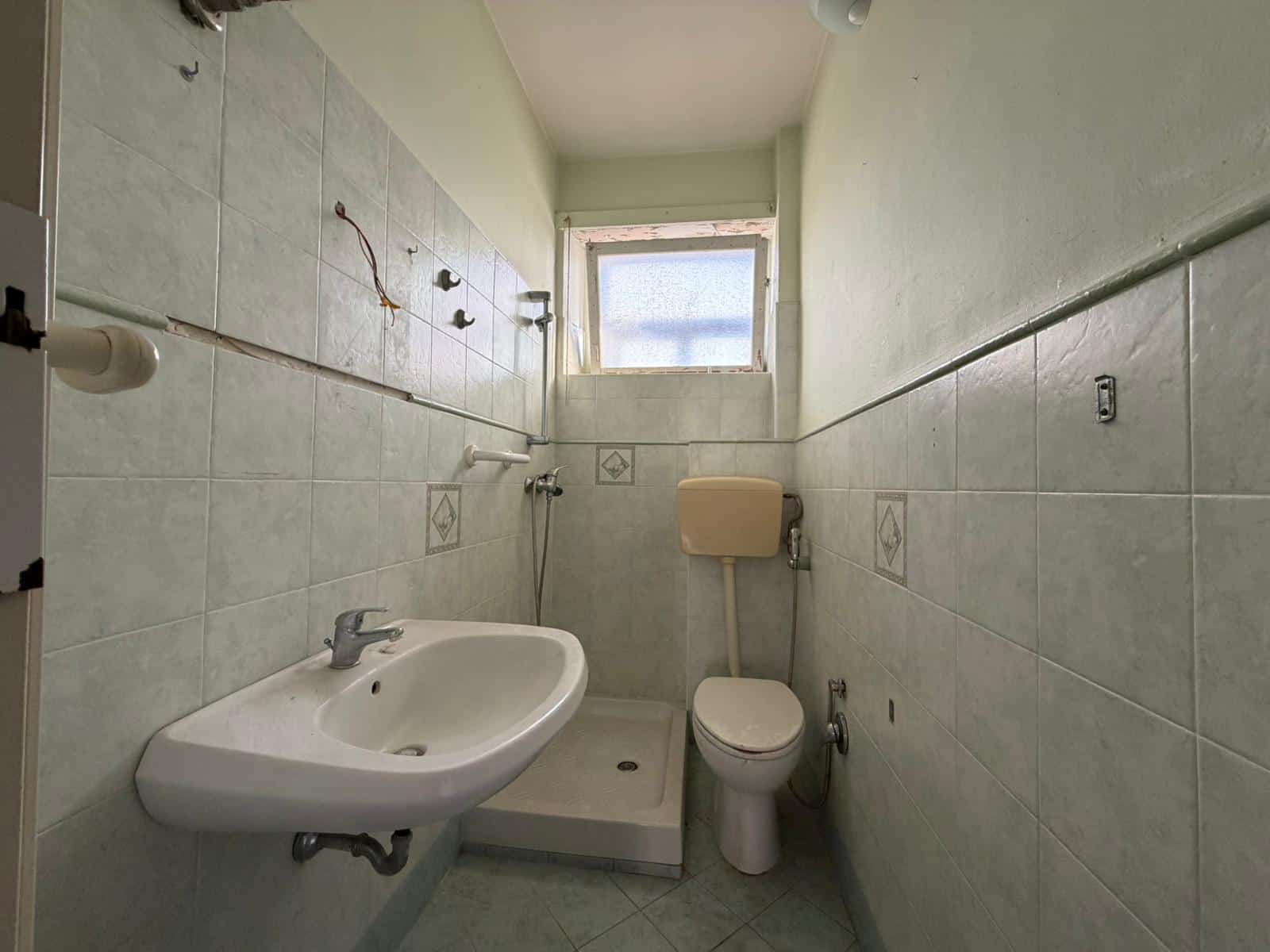 Lavandino e WC in un bagno piccolo con pareti rivestite in piastrelle chiare, finestra alta per ventilazione naturale, ambiente semplice e funzionale.
