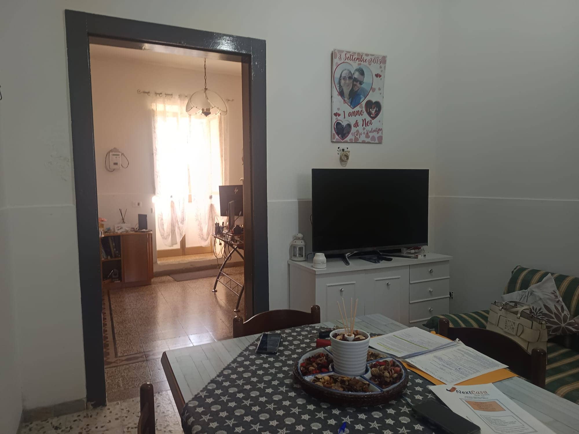 Interno soggiorno con tavolo, televisore e decorazioni, vista sulla stanza luminosa con finestra e tende leggere.