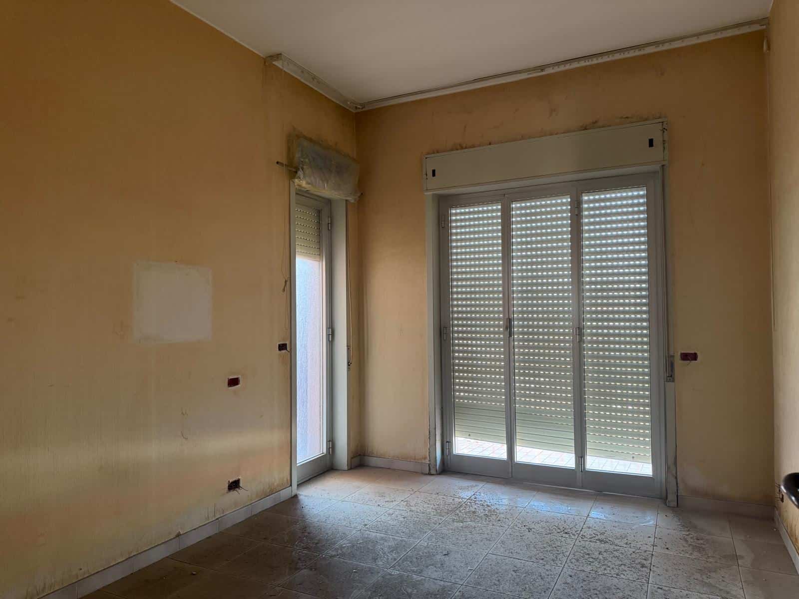 Stanza vuota in fase di ristrutturazione con pareti beige, pavimento in piastrelle e grandi finestre con tapparelle, pronta per essere rinnovata.