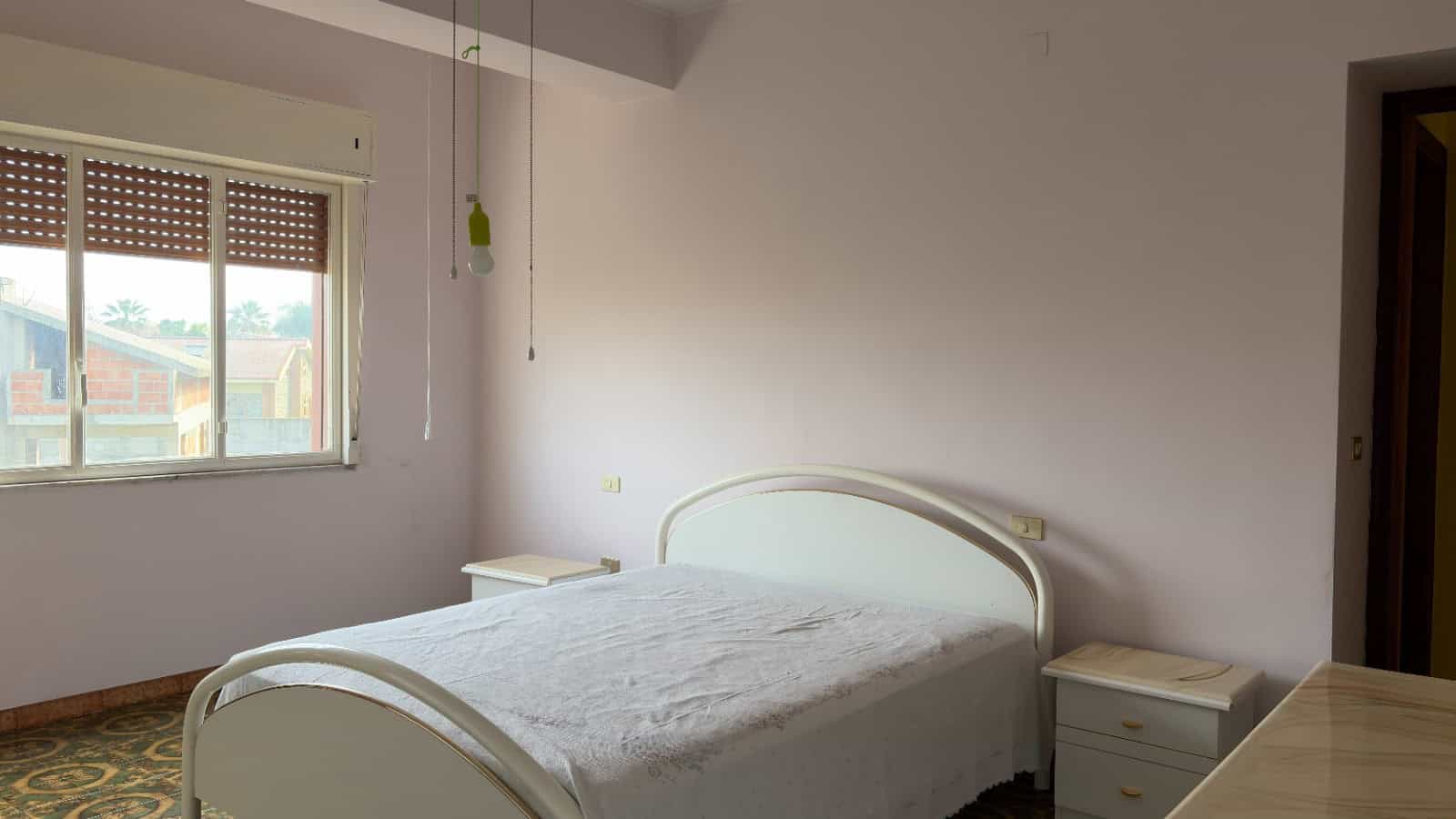 Ampio soggiorno con finestra, letto matrimoniale bianco e comodini in una casa moderna in Italia. Ideale per chi cerca soluzioni abitative confortevoli e funzionali.