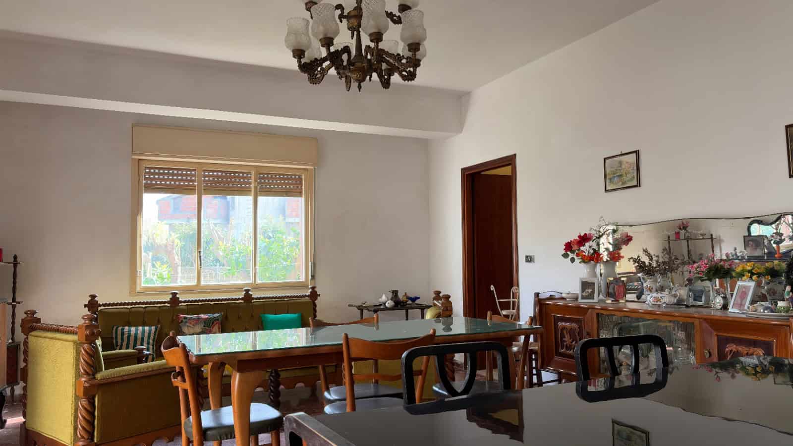 Ampio soggiorno con mobili d'epoca, tavolo in legno, divani e sedie, decorazioni floreali e specchio, illuminato da luce naturale attraverso una grande finestra, stile classico e accogliente.