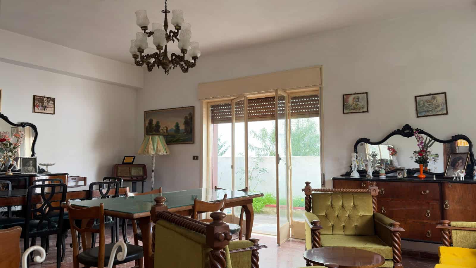 Ampia sala da pranzo con mobili classici, grande finestra con vista sul giardino, lampadario elegante e decorazioni vintage, ideale per vivere momenti conviviali in un ambiente accogliente e raffinato.