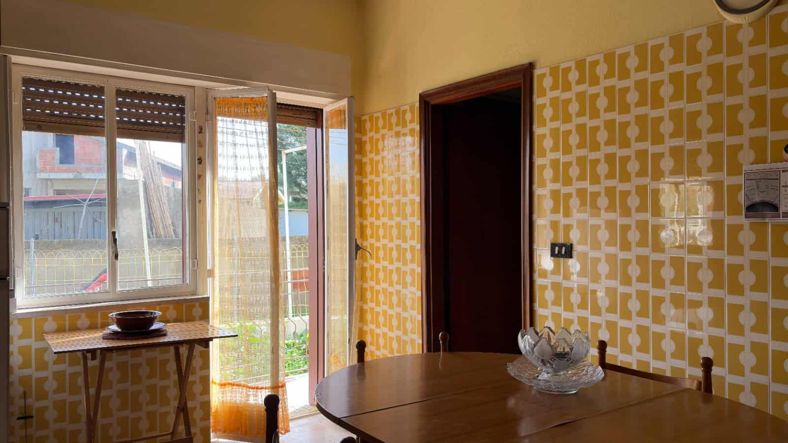 Ampia sala da pranzo con pareti gialle e motivo geometrico, grande finestra che permette l'ingresso di luce naturale, tavolo rotondo in legno con decorazioni eleganti, ideale per convivialità e moment.