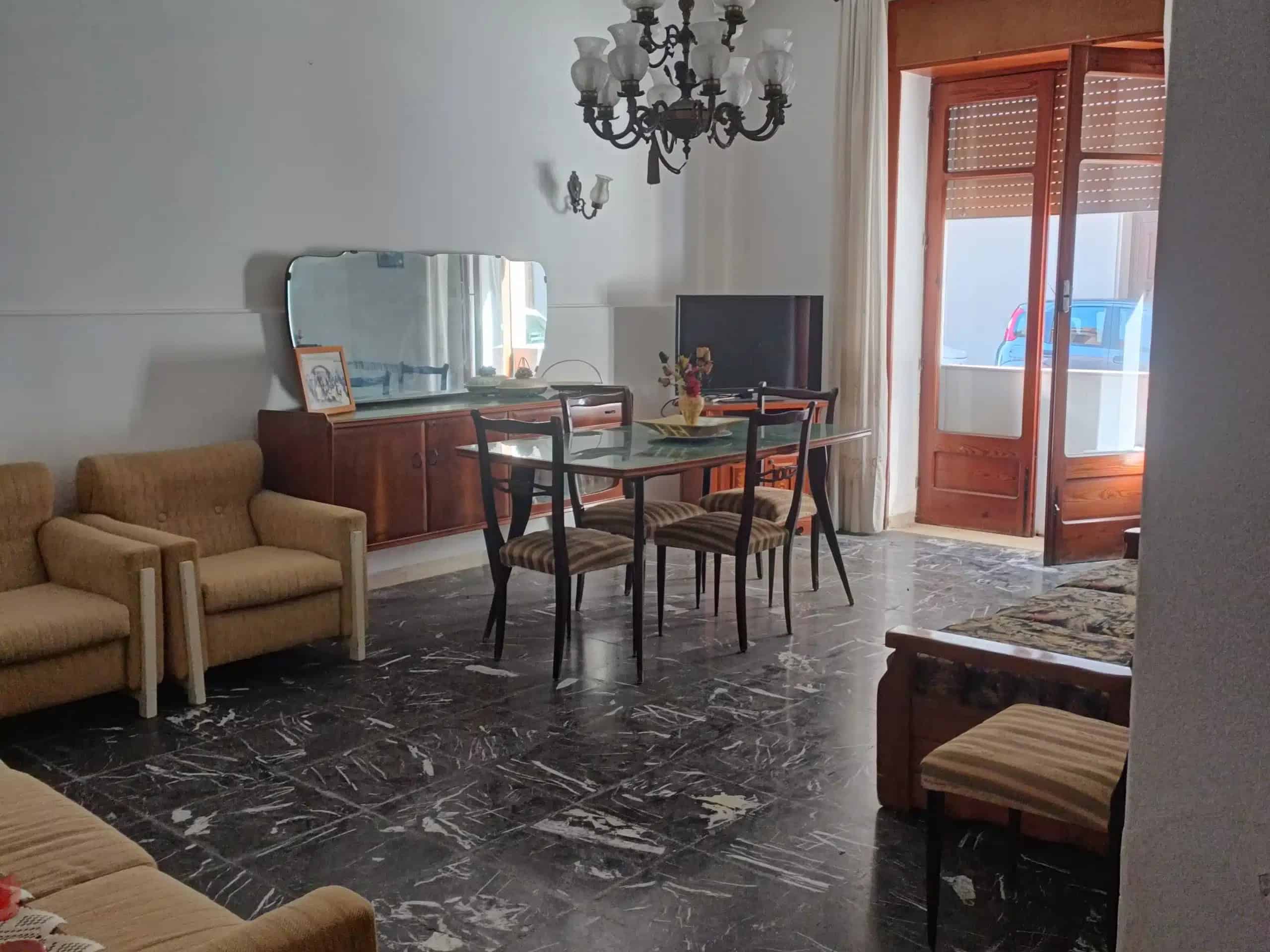 Il testo dell'immagine mostra un soggiorno luminoso con mobili classici, un tavolo da pranzo, sedie, un grande specchio, un televisore e un balcone con vista. Perfetto per ambienti di case in stile tr.