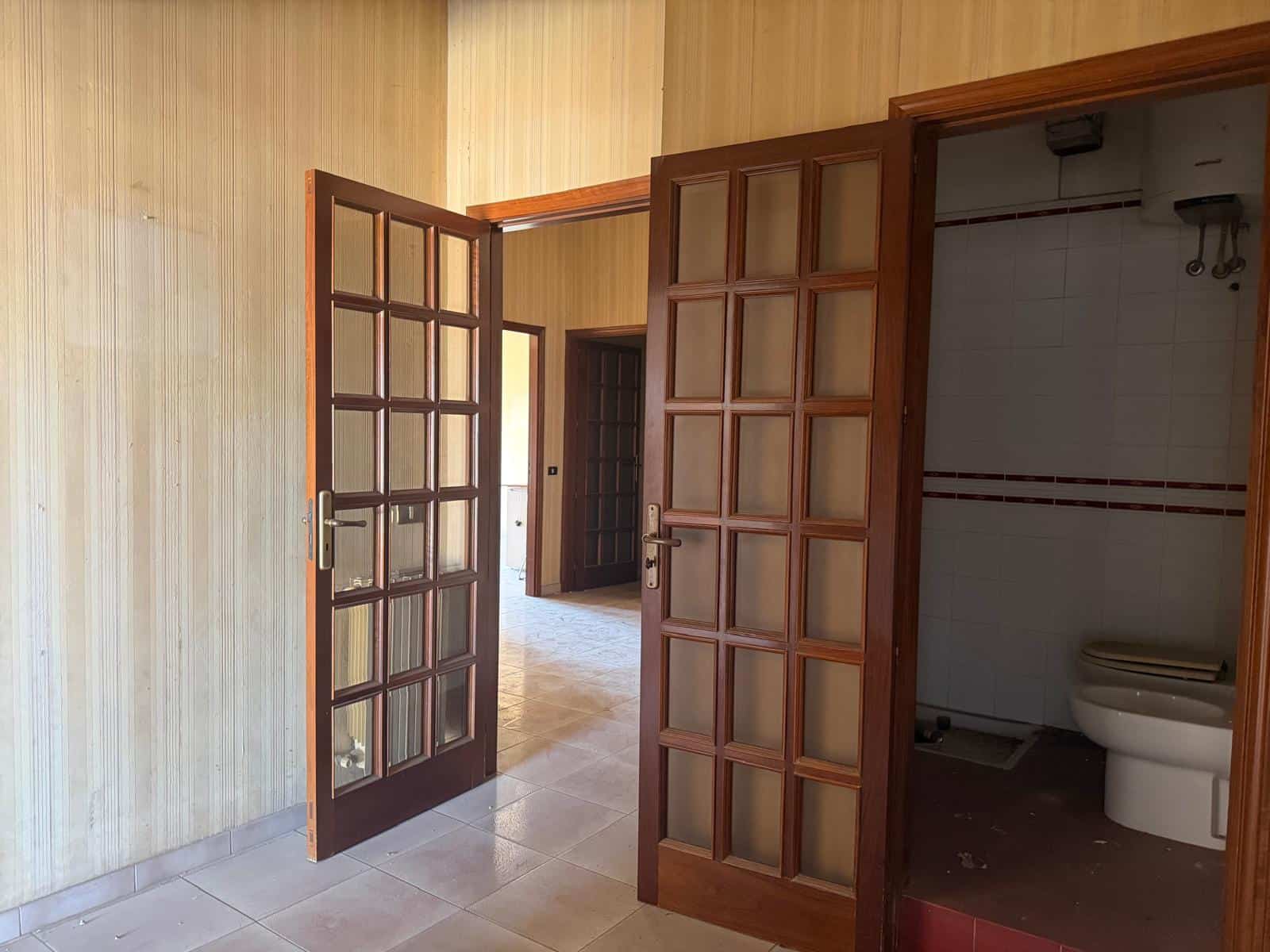 Ampia porta in legno con vetri a pannelli che conduce a un soggiorno luminoso e spazioso, con pareti rivestite in legno chiaro e pavimento in ceramica.