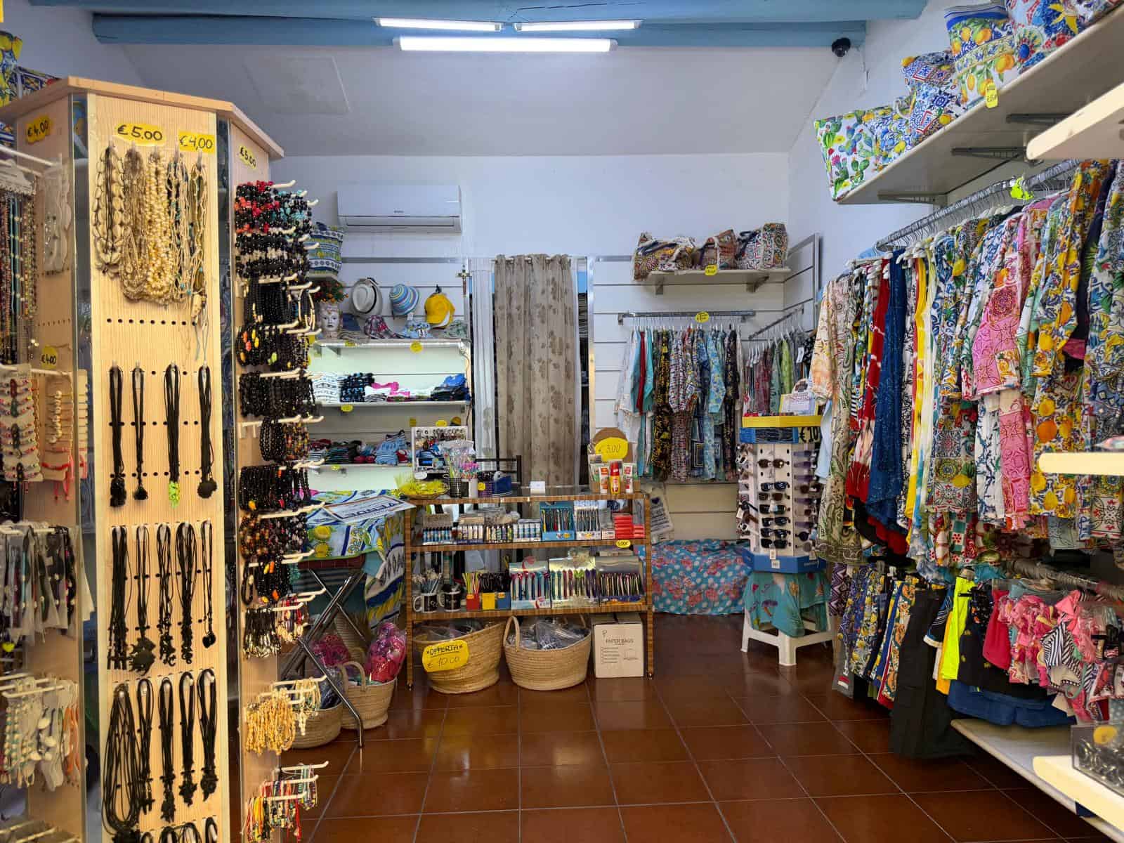 L'immagine mostra un negozio di souvenir e articoli di artigianato con abbigliamento colorato, accessori e decorazioni esposti su scaffali e pareti, ideale per acquisti di regali e ricordi locali.
