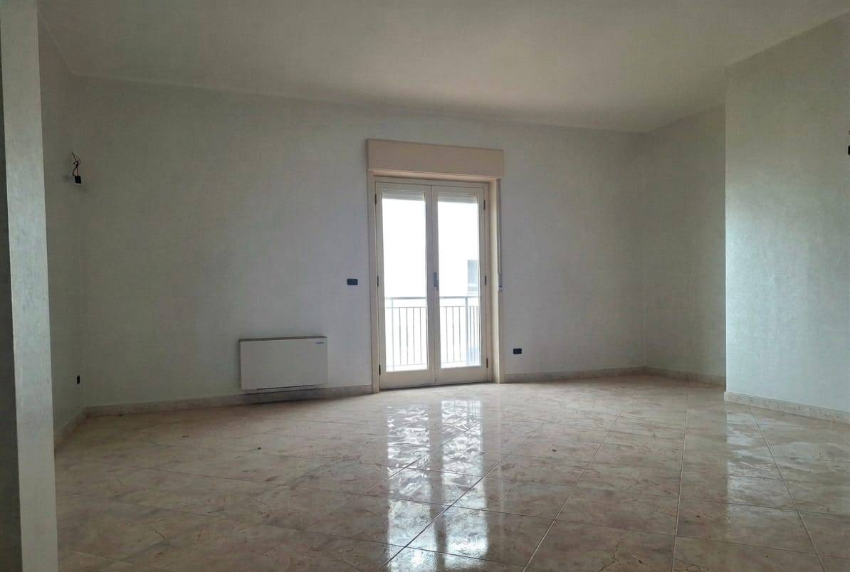 Ampia stanza vuota con pareti bianche, grande finestra con balcone che permette l'ingresso di luce naturale, pavimento in piastrelle beige, ideale per arredamento moderno e luminoso.