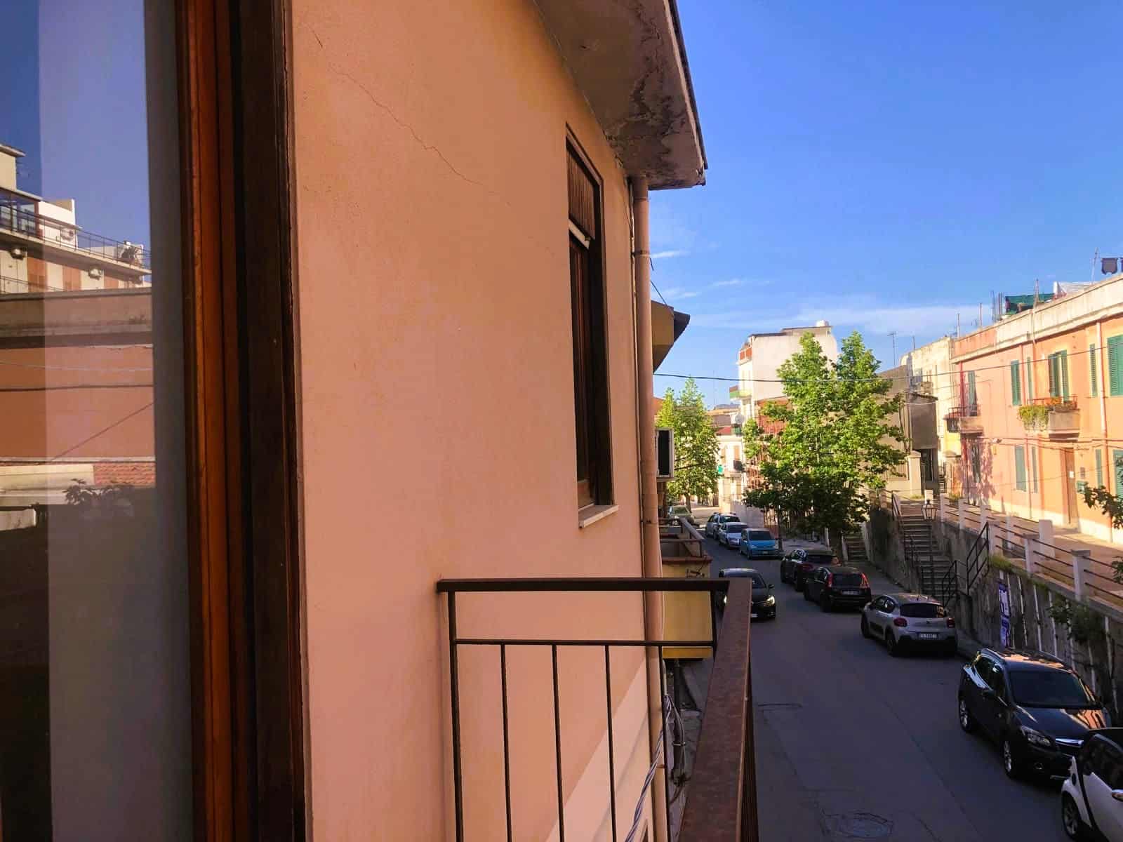 Vista di un balcone con ringhiera in metallo e strada urbana con auto e edifici sotto un cielo sereno, in un quartiere residenziale di città.