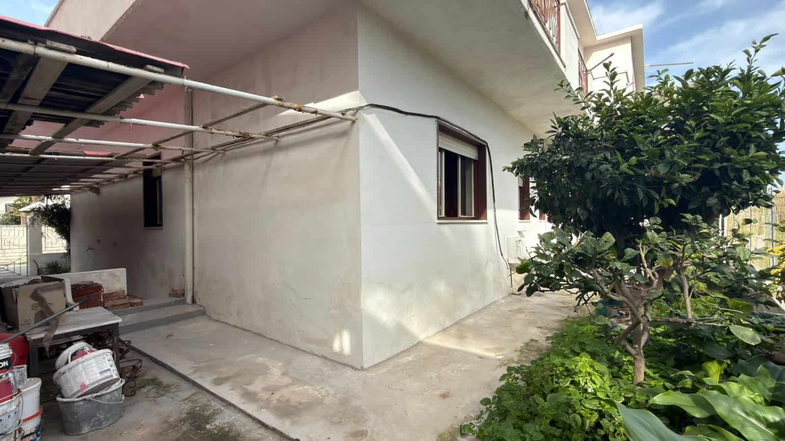 Ampia area esterna di una casa con pareti bianche, giardino con alberi e piante verdi, e spazio coperto per il parcheggio o attività all'aperto, ideale per vivere all'aperto e godere del verde.
