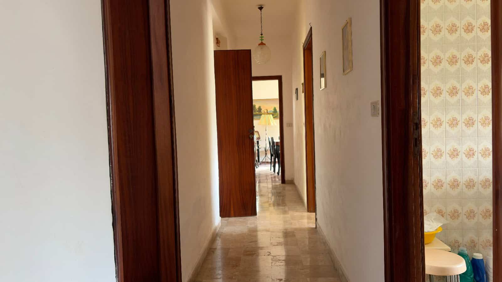 Ampia vista di un corridoio interno di una casa moderna, con pareti bianche e pavimento in marmo chiaro, illuminato da luce naturale e decorato con quadri alle pareti.