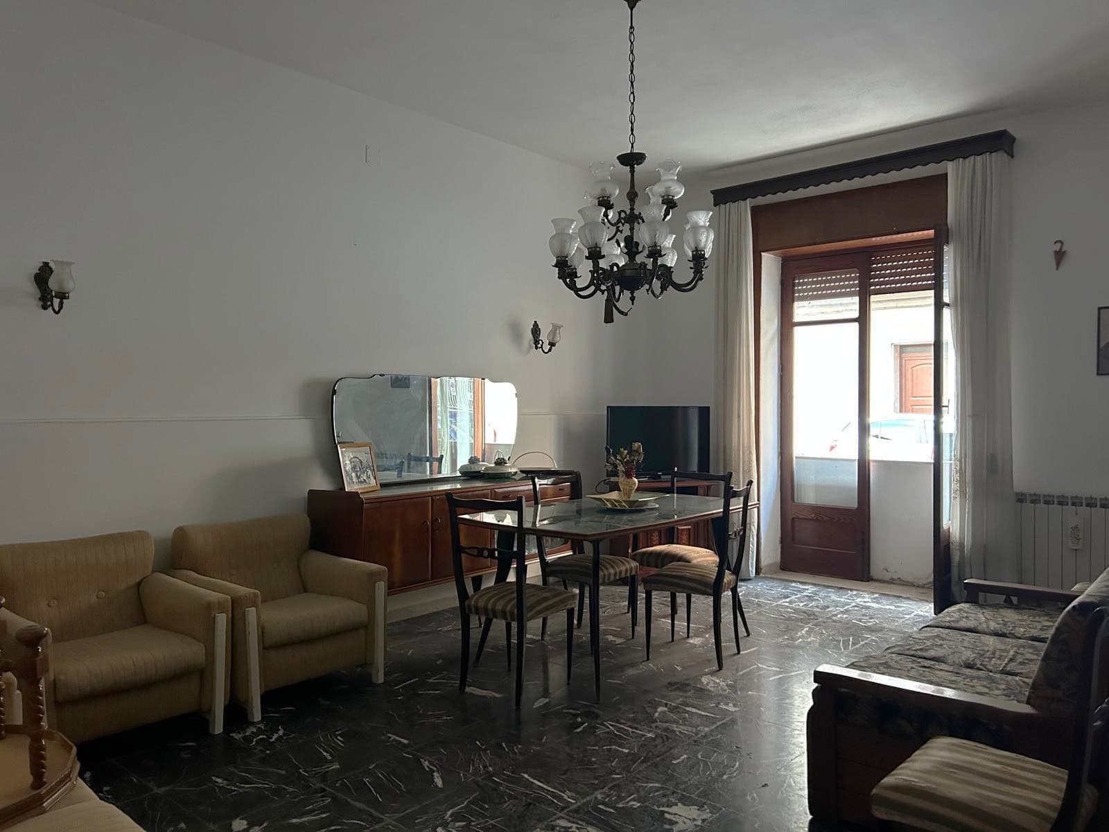 Interno di un soggiorno classico con mobili vintage, lampadario a chandelier e grande finestra con tende, arredamento elegante e atmosfera accogliente.