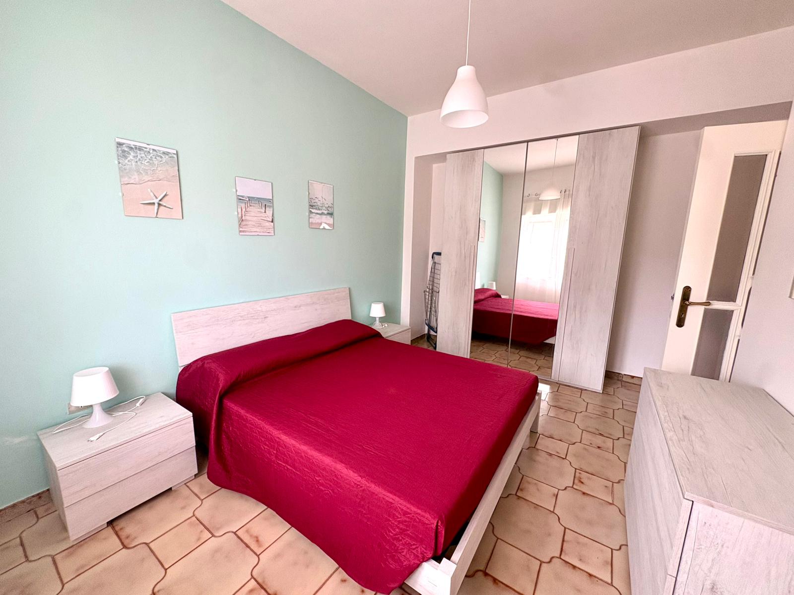 Una stanza da letto moderna con pareti verde menta, letto con copriletto rosso, due comodini con lampade e armadio a specchio, ambiente luminoso e accogliente.