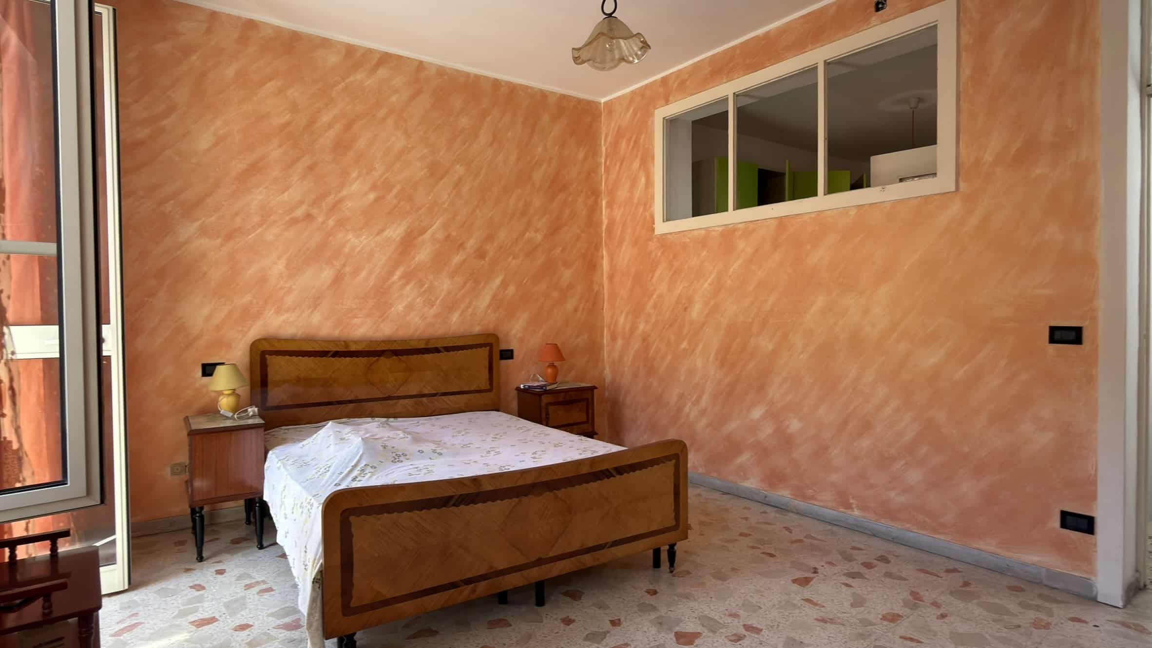 Letto matrimoniale in una camera da letto con pareti arancioni e pavimento in piastrelle, finestra grande che permette l'ingresso di luce naturale, arredamento semplice e accogliente.