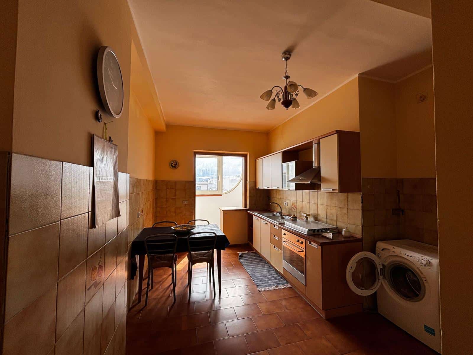 Una cucina luminosa e accogliente con mobili moderni, piano cottura e lavatrice, illuminata dalla luce naturale proveniente dalla finestra, ideale per la vita quotidiana e la preparazione dei pasti.