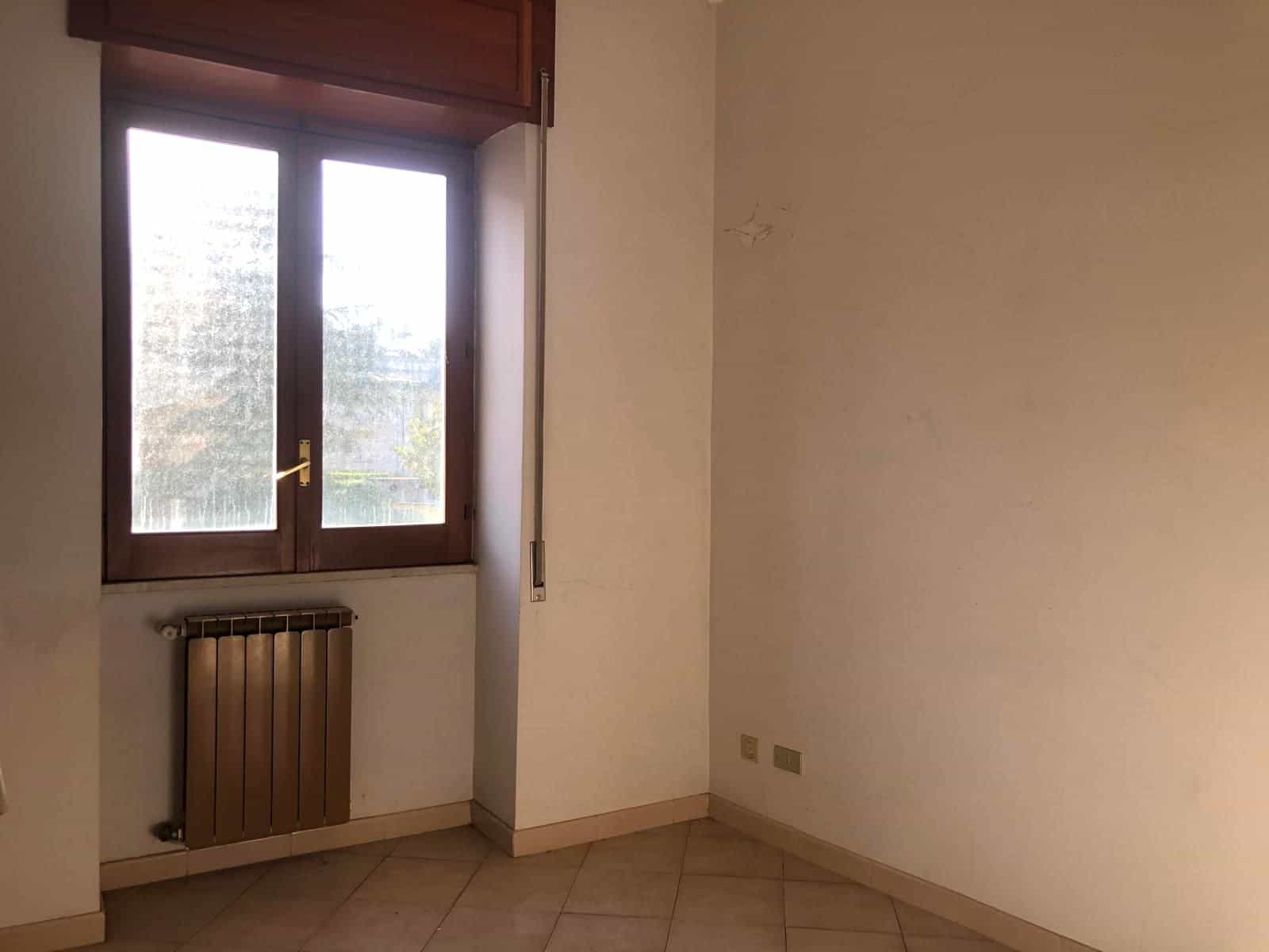 Stanza vuota con finestra e radiatore, pareti bianche e pavimento in piastrelle, ambiente luminoso e semplice.