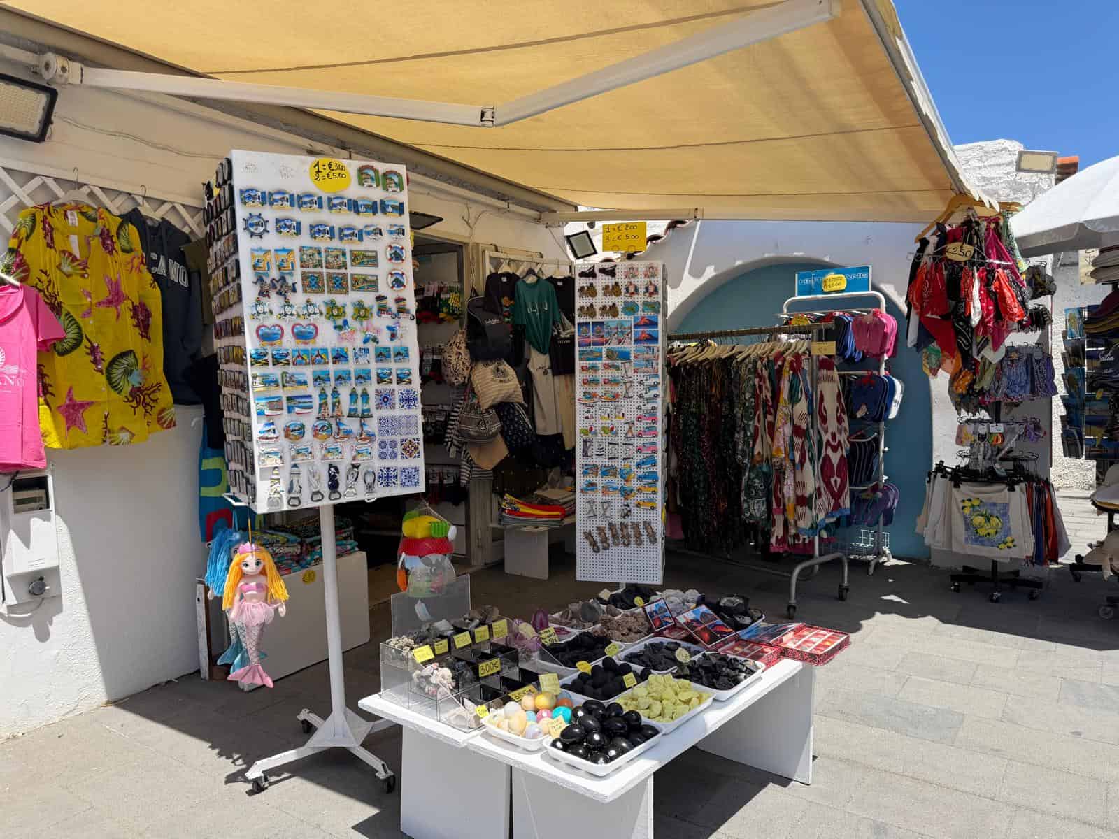 Pulsante di vendita di souvenir e accessori moda all'aperto con abbigliamento colorato e gadget vari, situato in una zona di mercato soleggiata con tende e strutture di supporto.