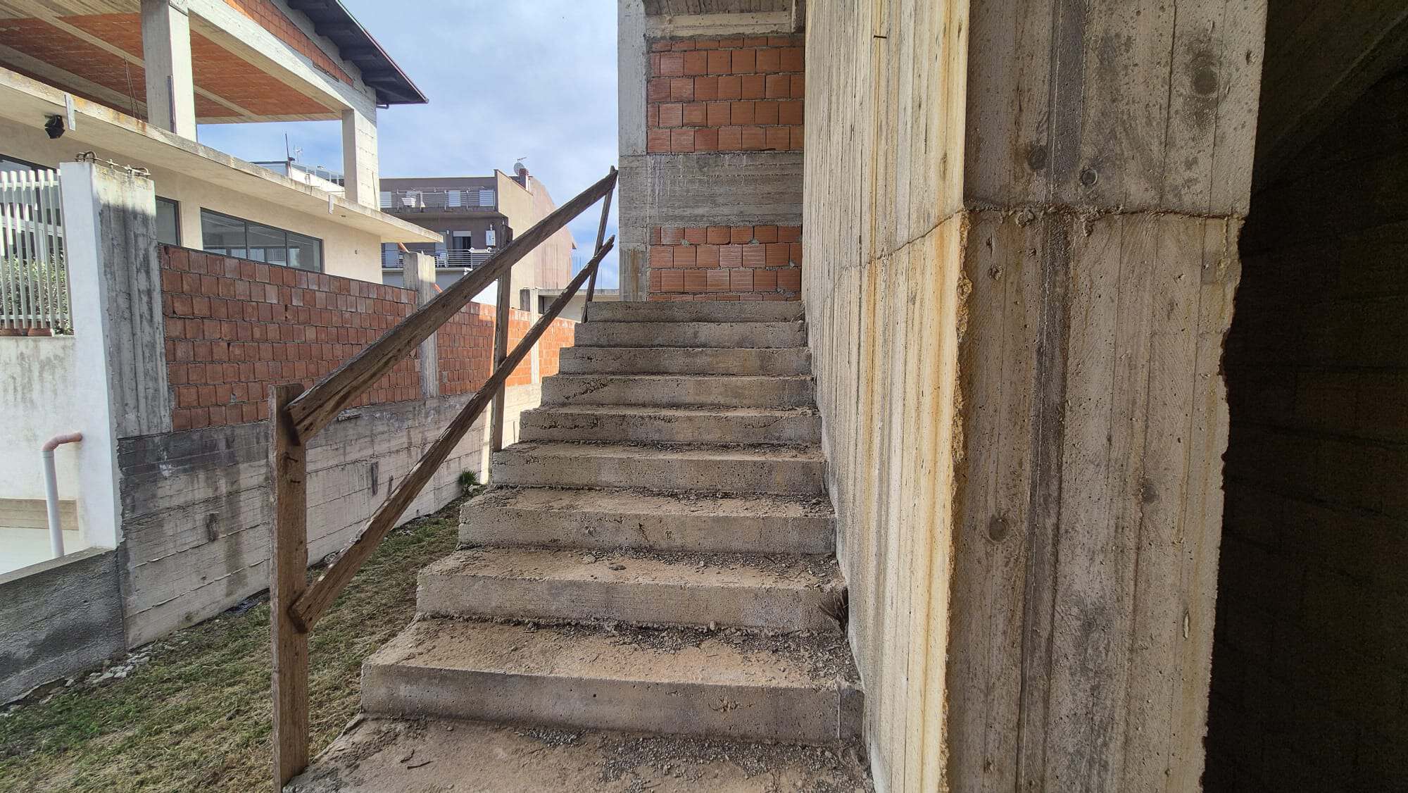 Scale esterna in cemento con ringhiera in legno, parte di un edificio in costruzione, con sfondo di case moderne e cielo nuvoloso.