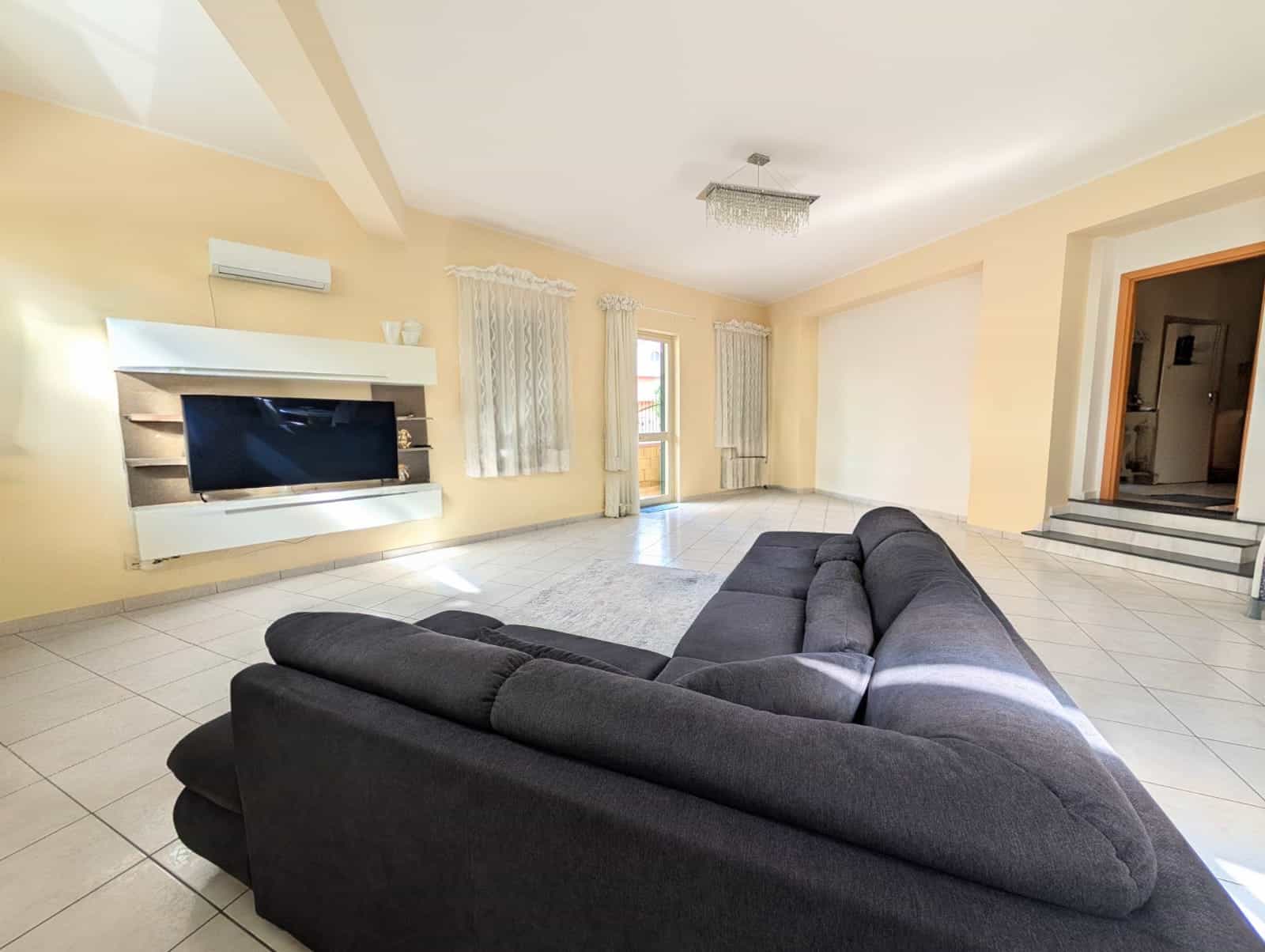 Ampia zona living moderna con divano nero, parete TV e grandi finestre con tende leggere, luminoso e accogliente, ideale per relax e intrattenimento in casa.