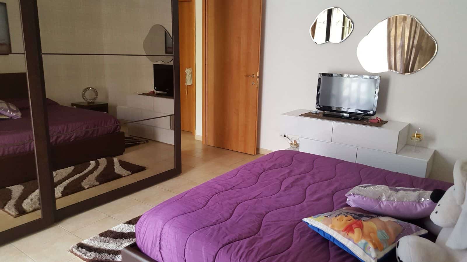 Letto matrimoniale con biancheria viola, specchi decorativi e televisore in camera da letto moderna.