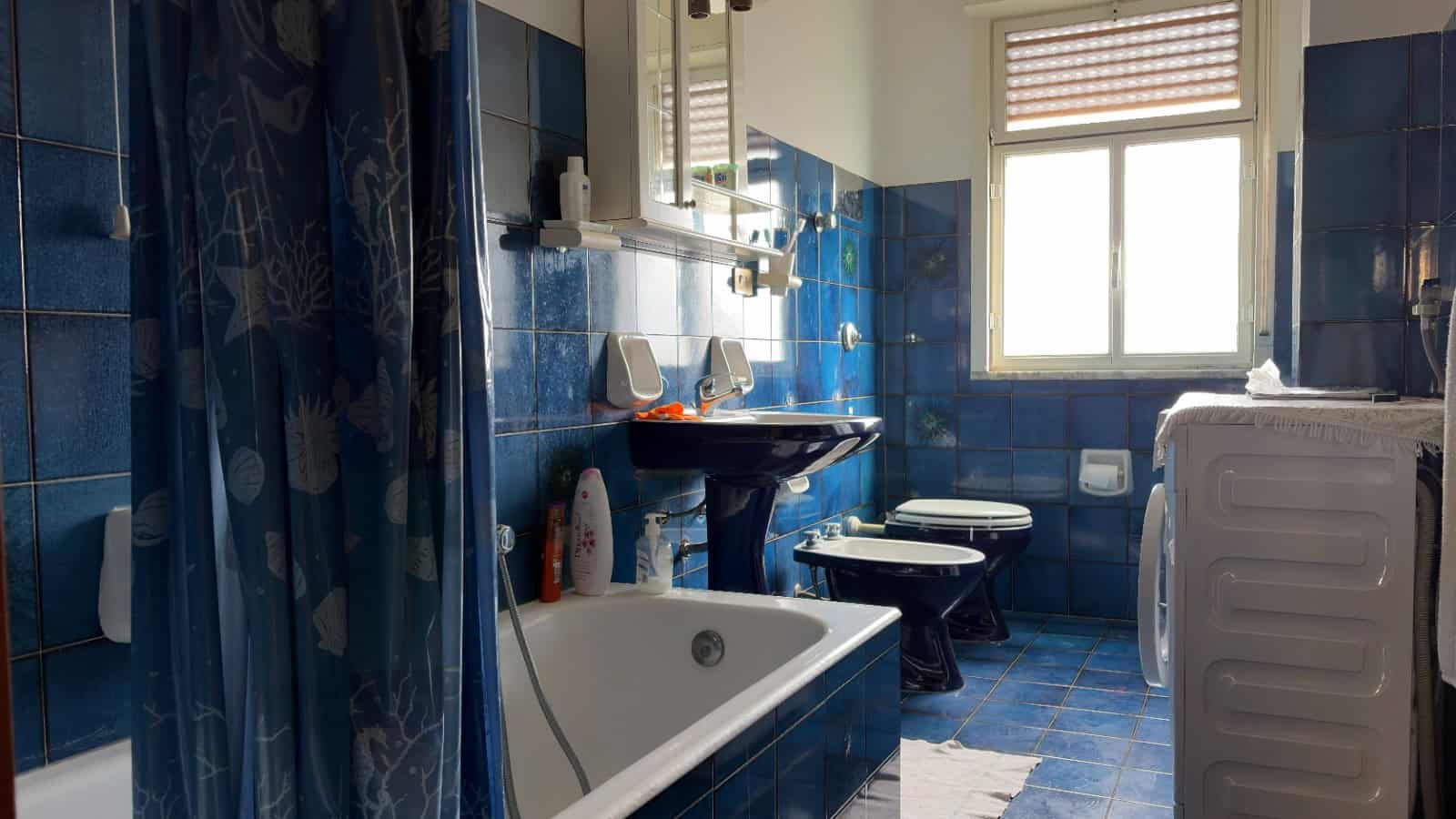 Doccia e vasca da bagno in un bagno con piastrelle blu, lavabo e bidet, finestra che permette l'ingresso di luce naturale, ambiente pulito e funzionale, ideale per il comfort quotidiano.