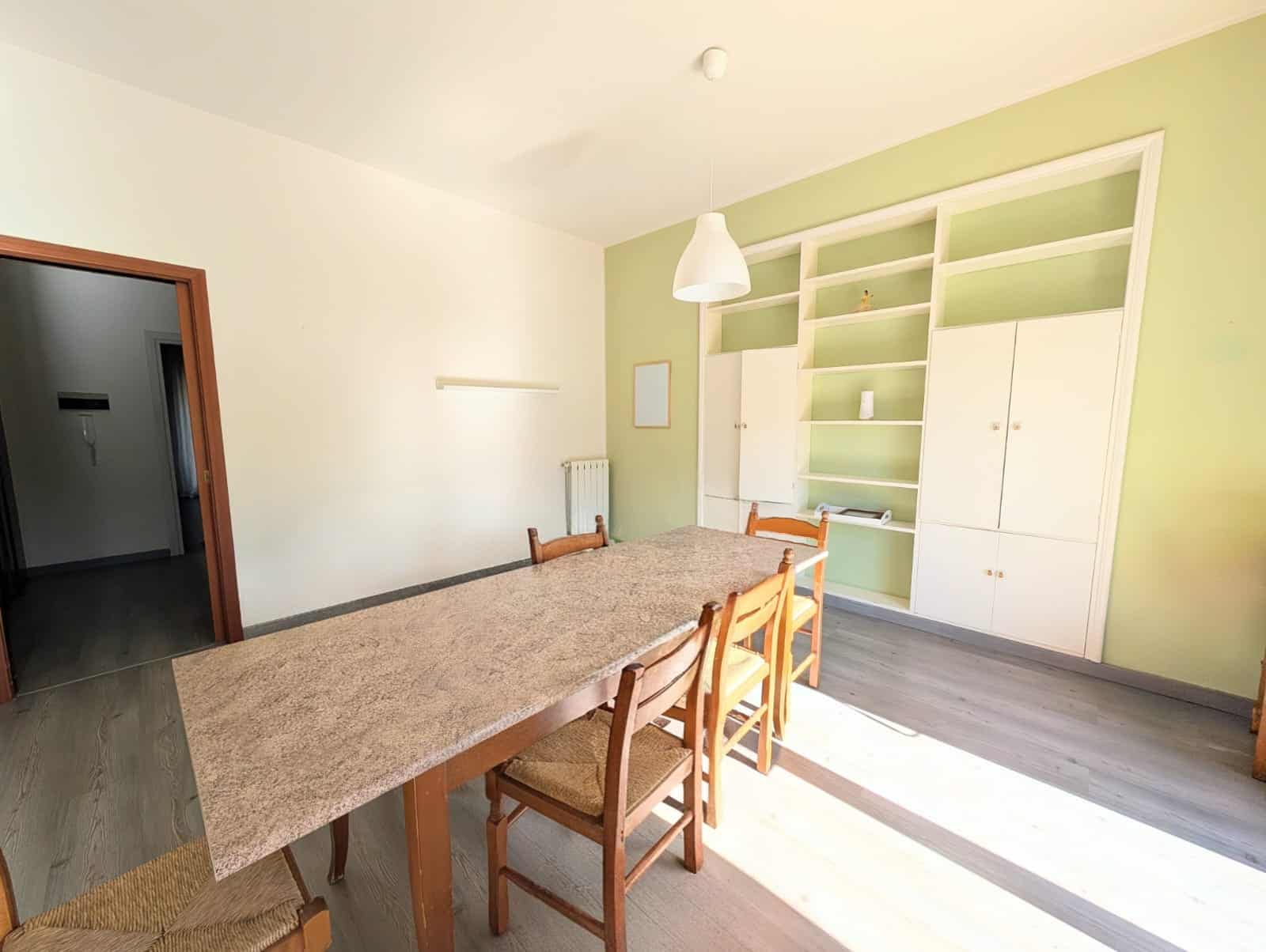 Ampia sala da pranzo con tavolo in marmo e sedie in legno, pareti verde chiaro e libreria bianca con armadi, illuminata da lampadario sospeso, ambiente luminoso e accogliente.