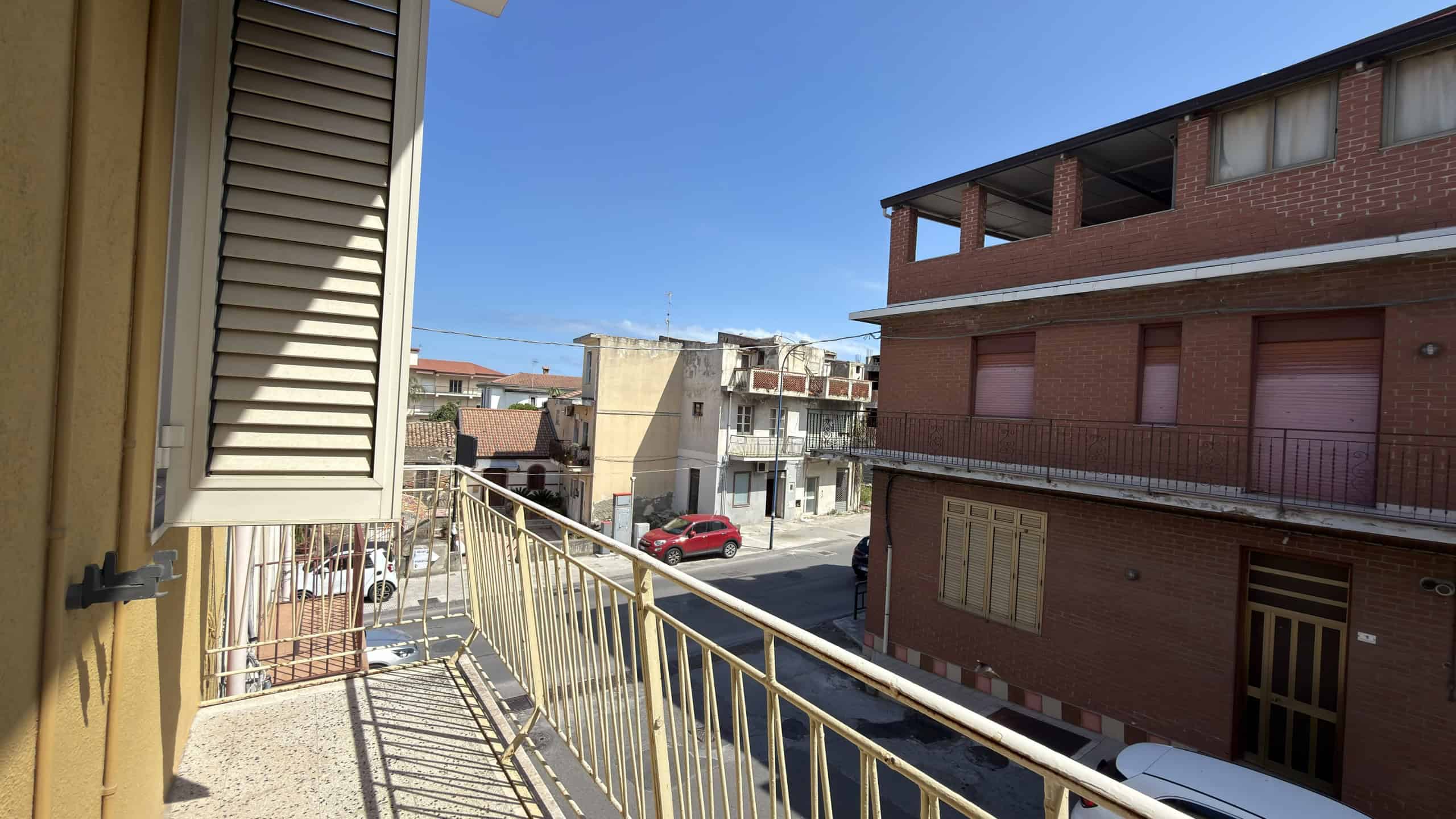 Ampio balcone con vista su strada urbana, con edifici residenziali in mattoni e auto parcheggiate, sotto un cielo sereno, in un quartiere residenziale di città.