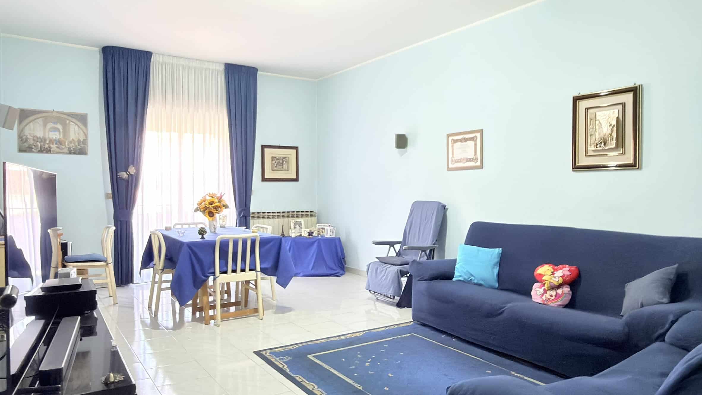 Una sala soggiorno luminosa con pareti azzurre, divano blu, tavolo da pranzo con tovaglia blu, sedie bianche e decorazioni floreali, creando un ambiente accogliente e elegante.