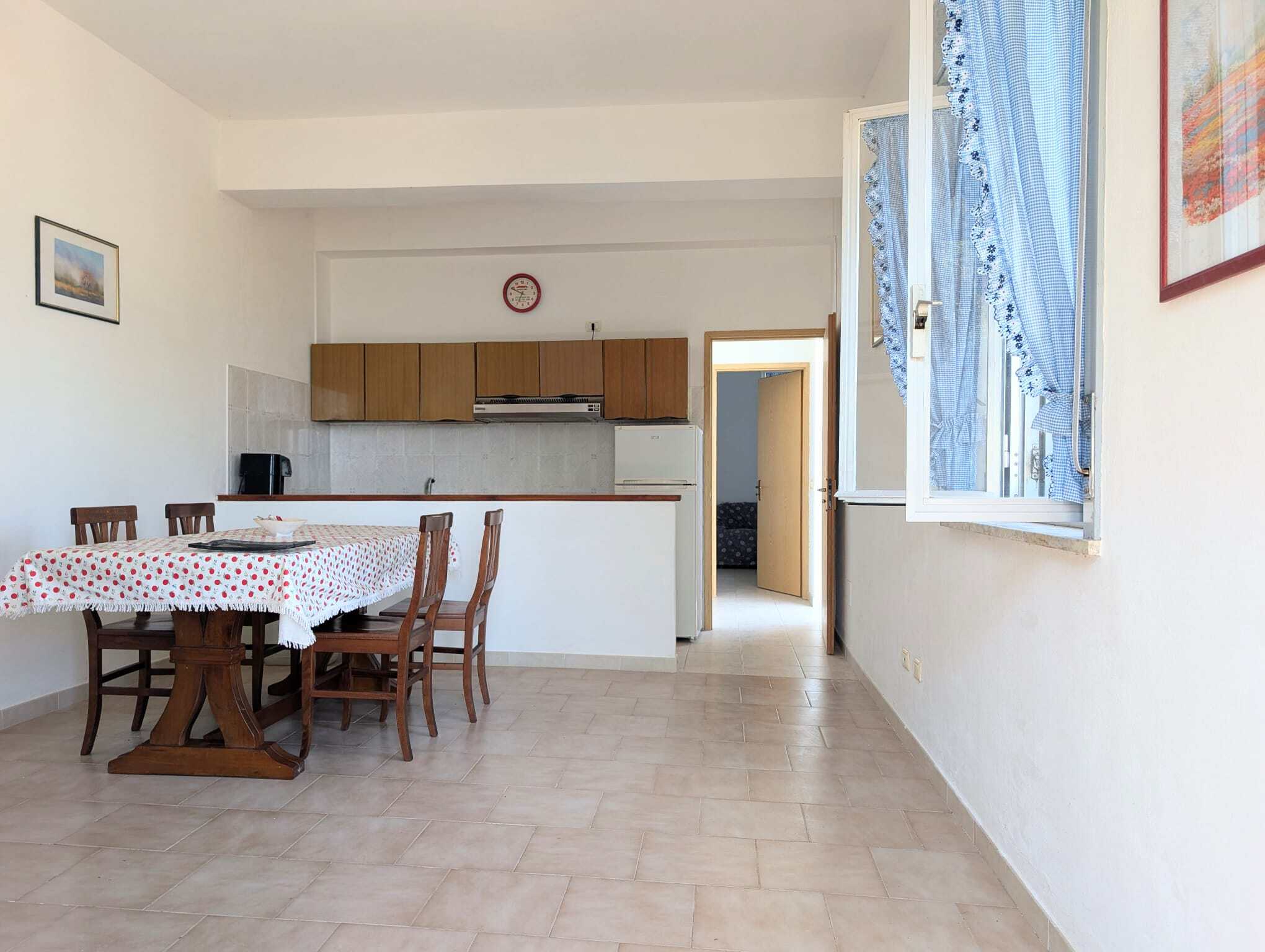 Ampia zona pranzo con tavolo e sedie in legno, cucina aperta e luminosa con finestra che permette l'ingresso di luce naturale, pareti bianche e pavimento in piastrelle beige, ambiente accogliente e fu.