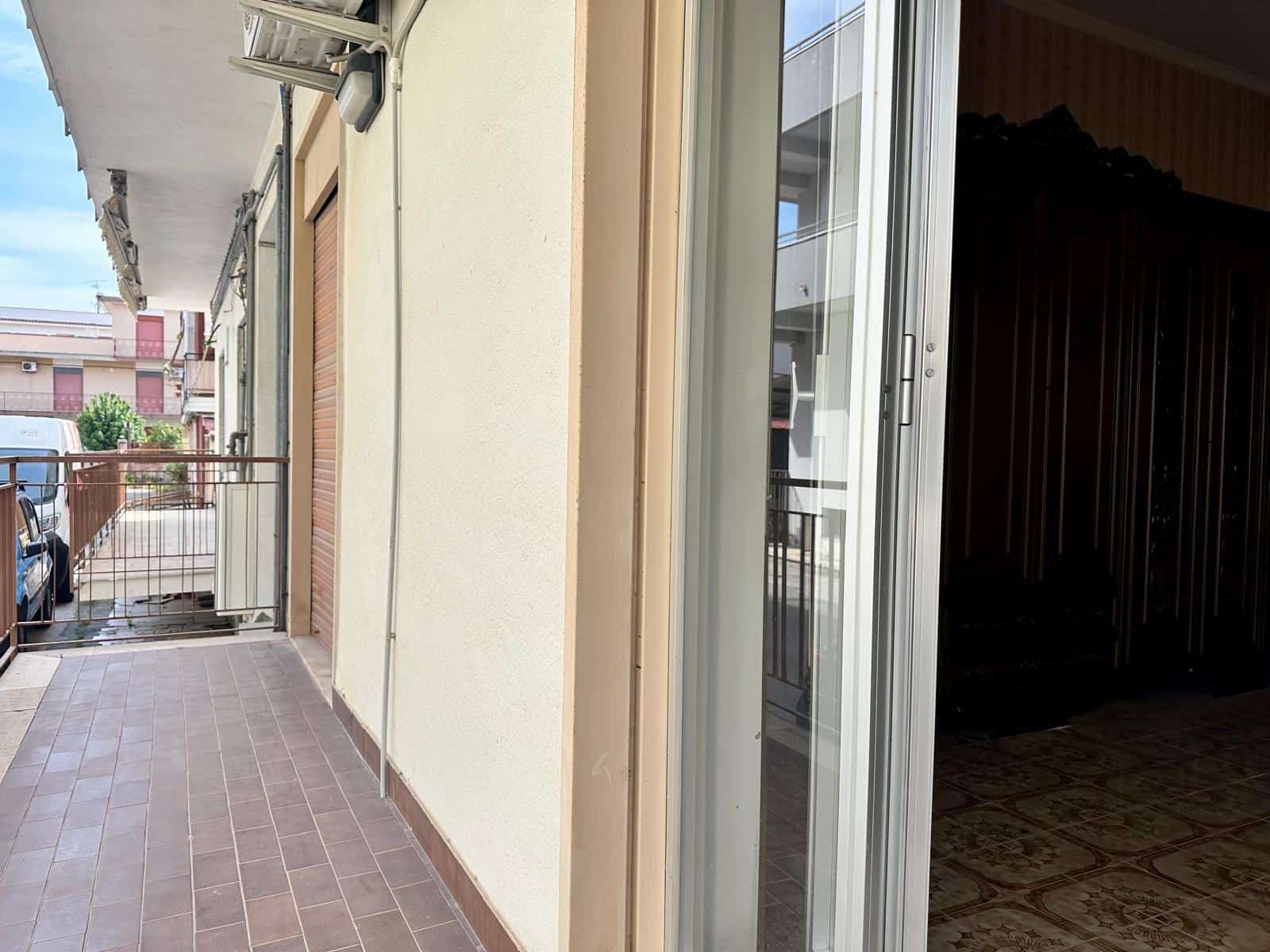 Balcone di un appartamento con vista sulla strada, pavimentato in piastrelle di ceramica, con ringhiera in metallo e porta scorrevole in vetro, in un contesto urbano residenziale.