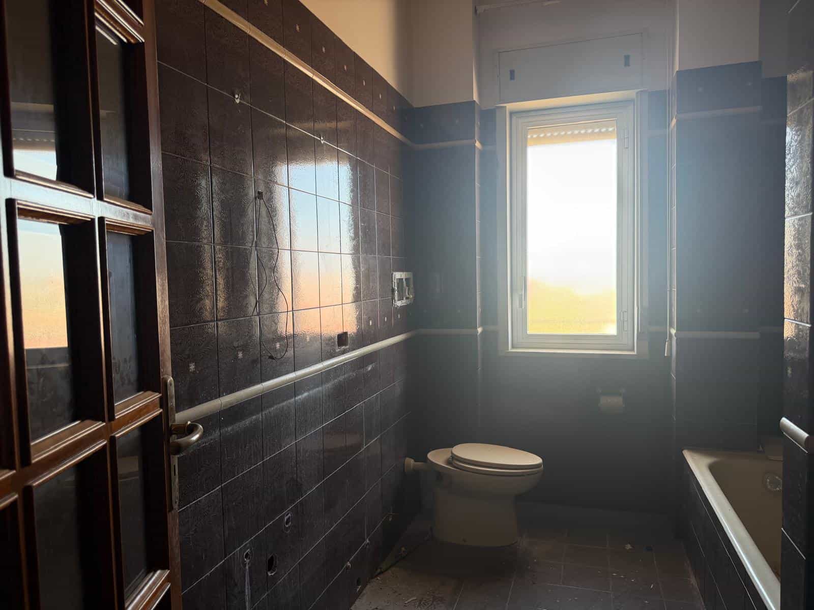 Bagno moderno con pareti rivestite in piastrelle scure, finestra luminosa che permette l'ingresso di luce naturale, lavabo e vasca da bagno, ideale per ambienti di comfort e stile.