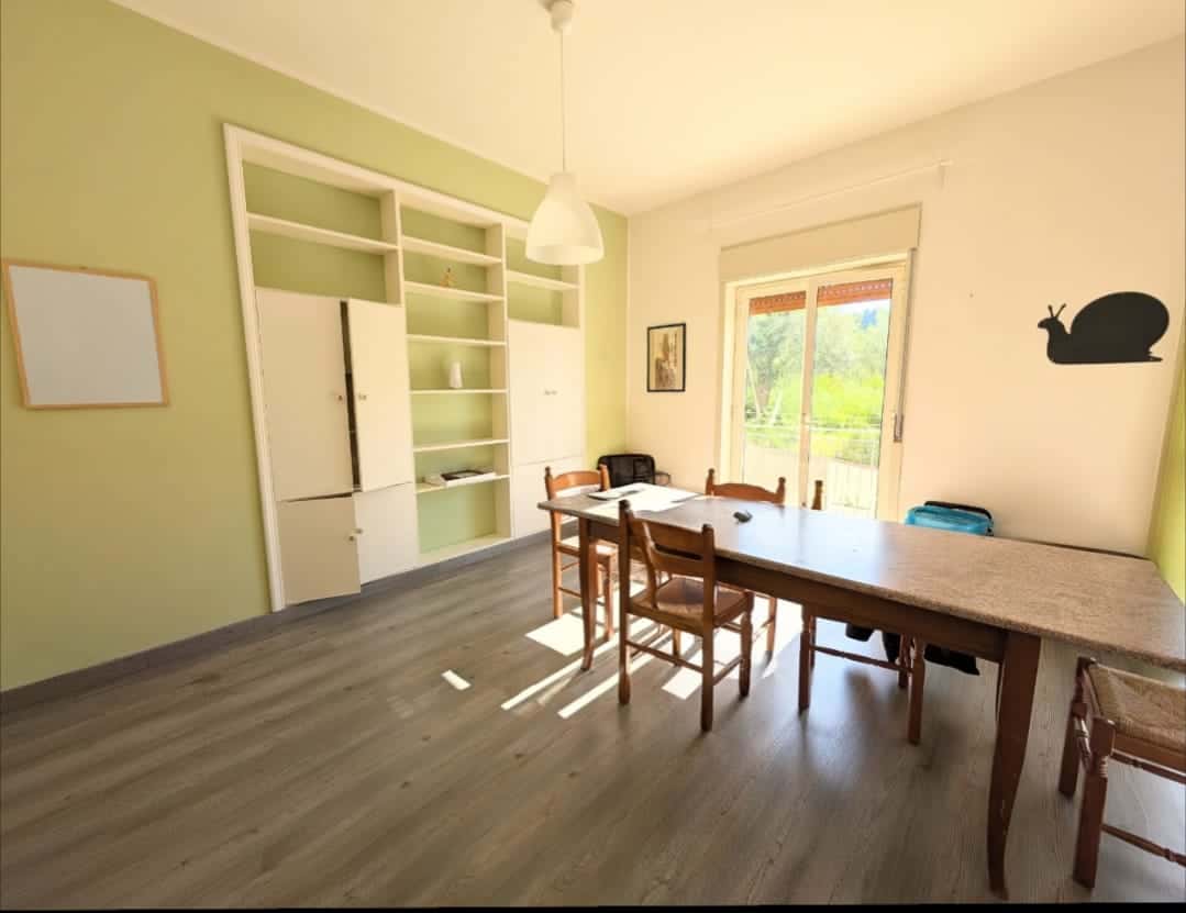 Ampia sala da pranzo luminosa con pareti verde chiaro, grande tavolo in legno e sedie, finestra che lascia entrare molta luce naturale, decorazioni minimaliste e ambiente accogliente.