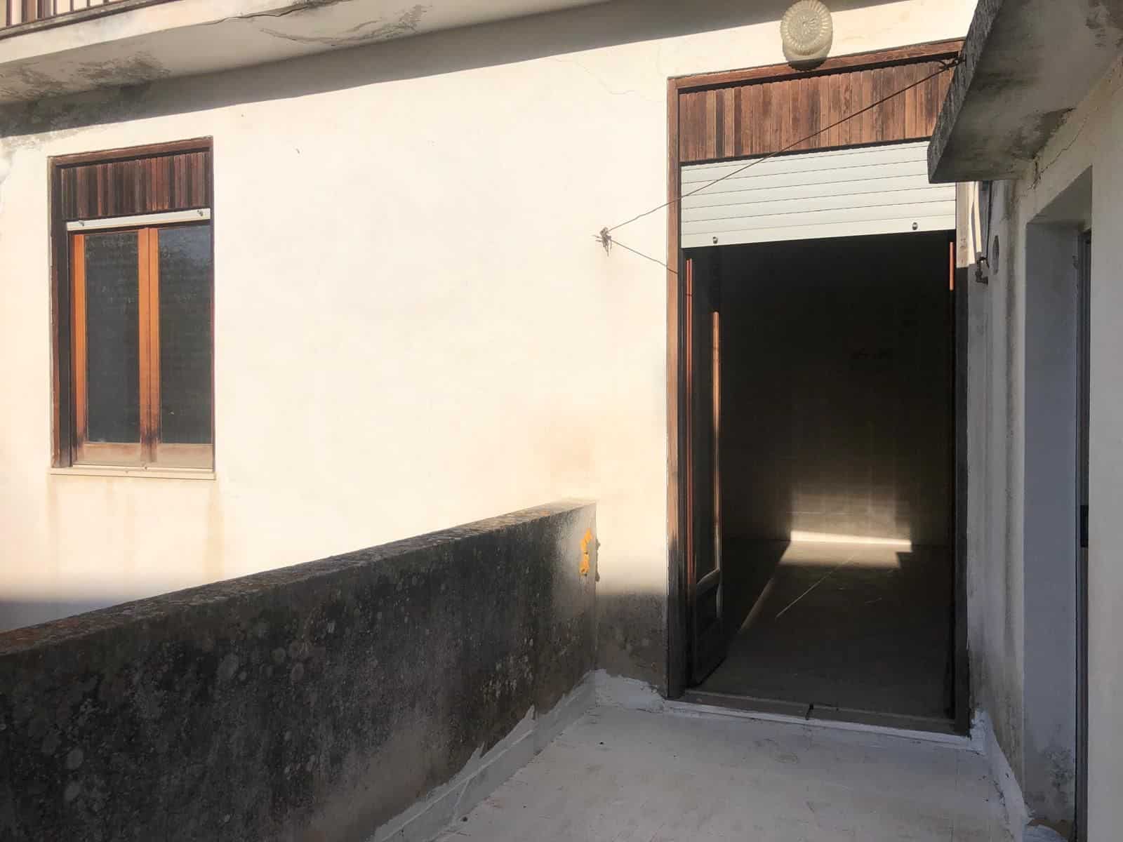 Ampio ingresso di una casa con porta scorrevole in legno e finestra con persiana, facciata esterna in stile rustico, illuminata dal sole, con muro di cinta e pavimentazione in cemento.