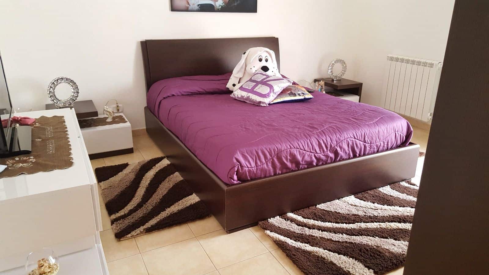 Letto matrimoniale con coperta viola e cuscino decorativo, in una camera moderna e accogliente.