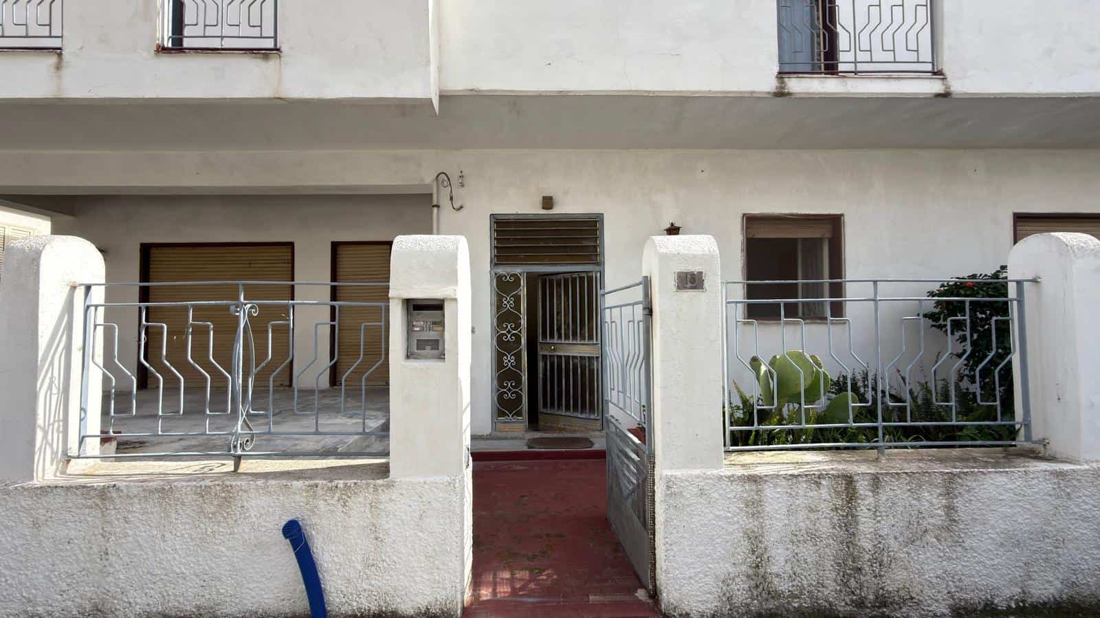 Immagine di una casa con ingresso pedonale e recinzione in metallo, facciata bianca e finestre con grate in ferro, ambiente residenziale tranquillo e ben curato.