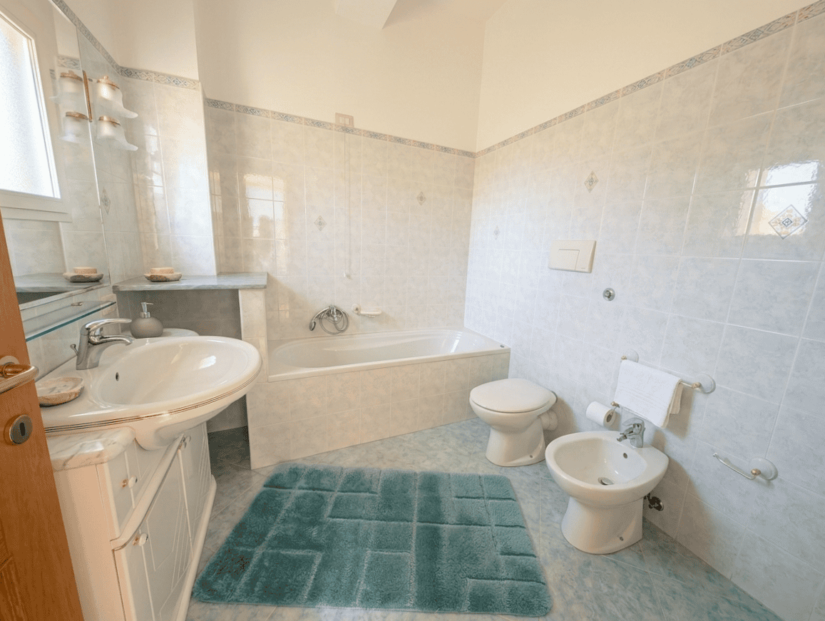 Lavabo, bidet e vasca da bagno in una stanza luminosa con pareti rivestite di piastrelle chiare e pavimento in ceramica azzurra, arredamento semplice e funzionale.