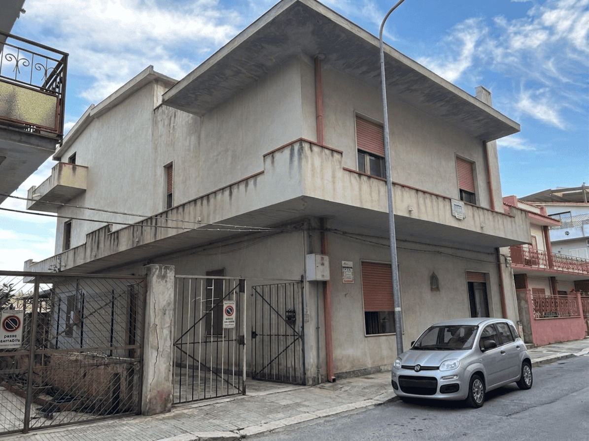 Ampia casa residenziale in cemento con balcone e garage, situata in zona urbana, con auto parcheggiata davanti e cielo sereno sullo sfondo.