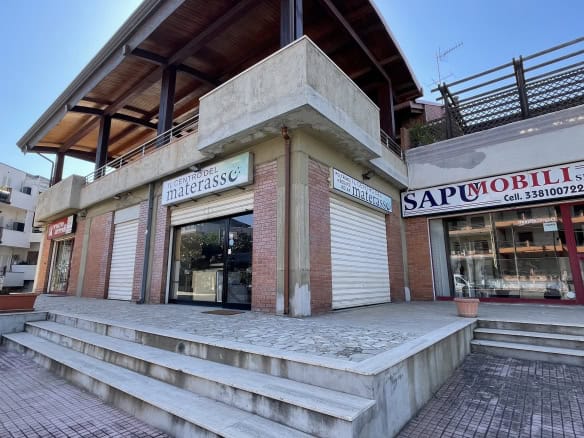 Pannello frontale di un edificio commerciale con negozi in vendita o affitto, con ampie vetrine e accesso tramite scalinate, situato in un'area urbana moderna e ben sviluppata.