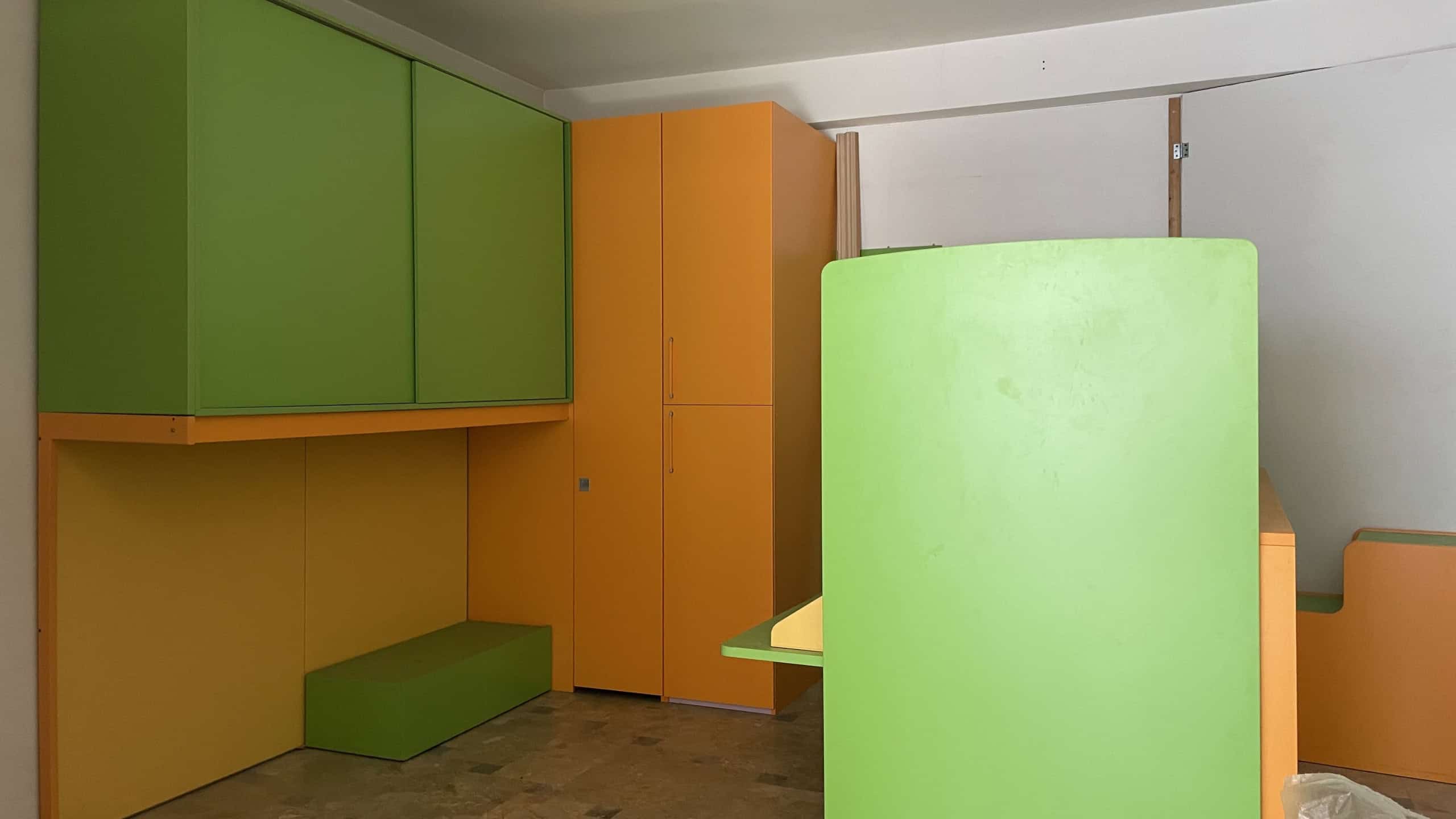 Armadio a muro colorato in soggiorno con mobili modulari in verde, arancione e giallo, ideale per arredare con stile e funzionalità. Soluzione di arredo moderna e vivace per la casa.