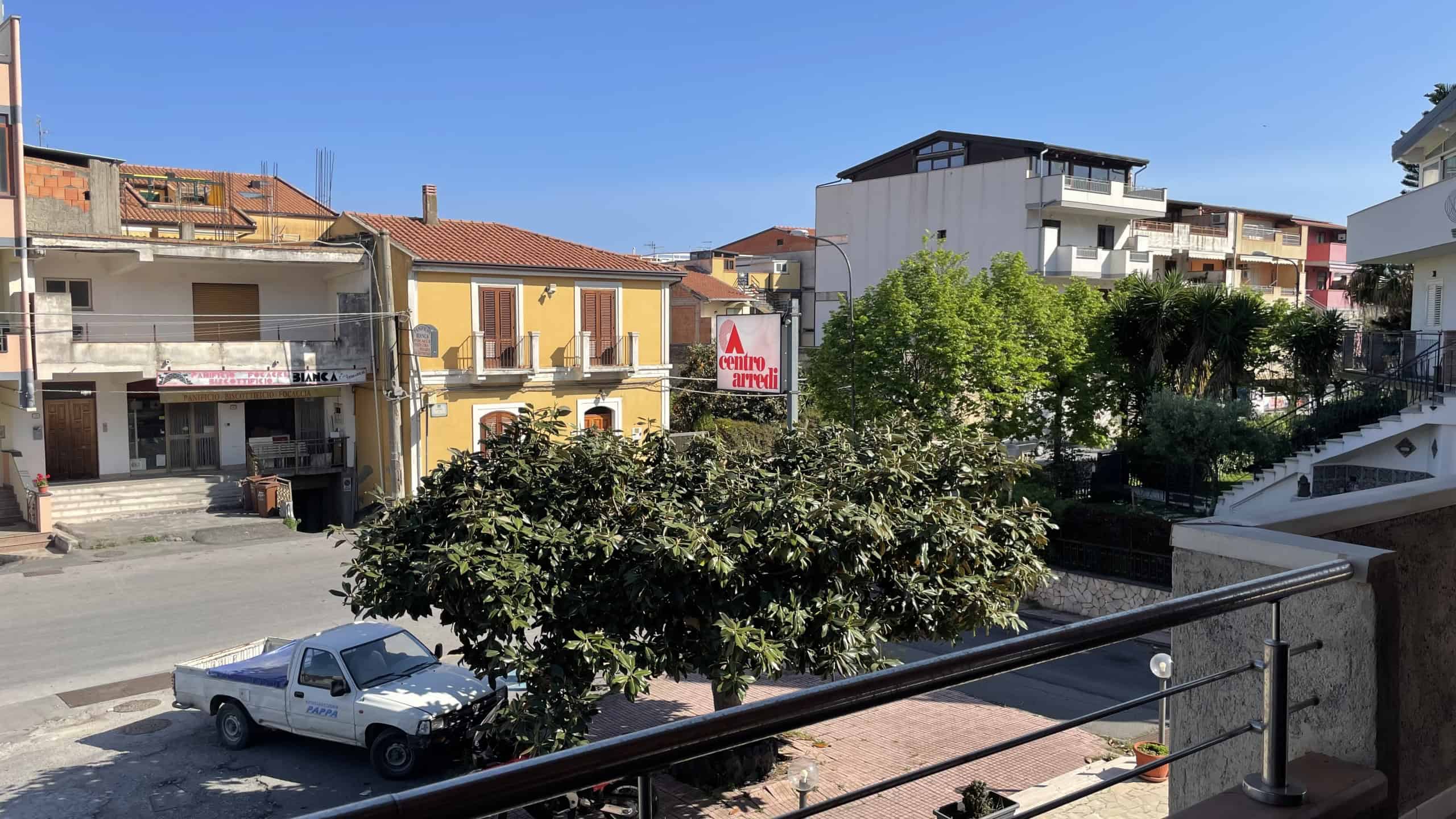 Piante verdi e strada in un quartiere residenziale con edifici colorati e moderni, cielo sereno, atmosfera tranquilla e vivace, ideale per acquisto o affitto di immobili a NextCasa.