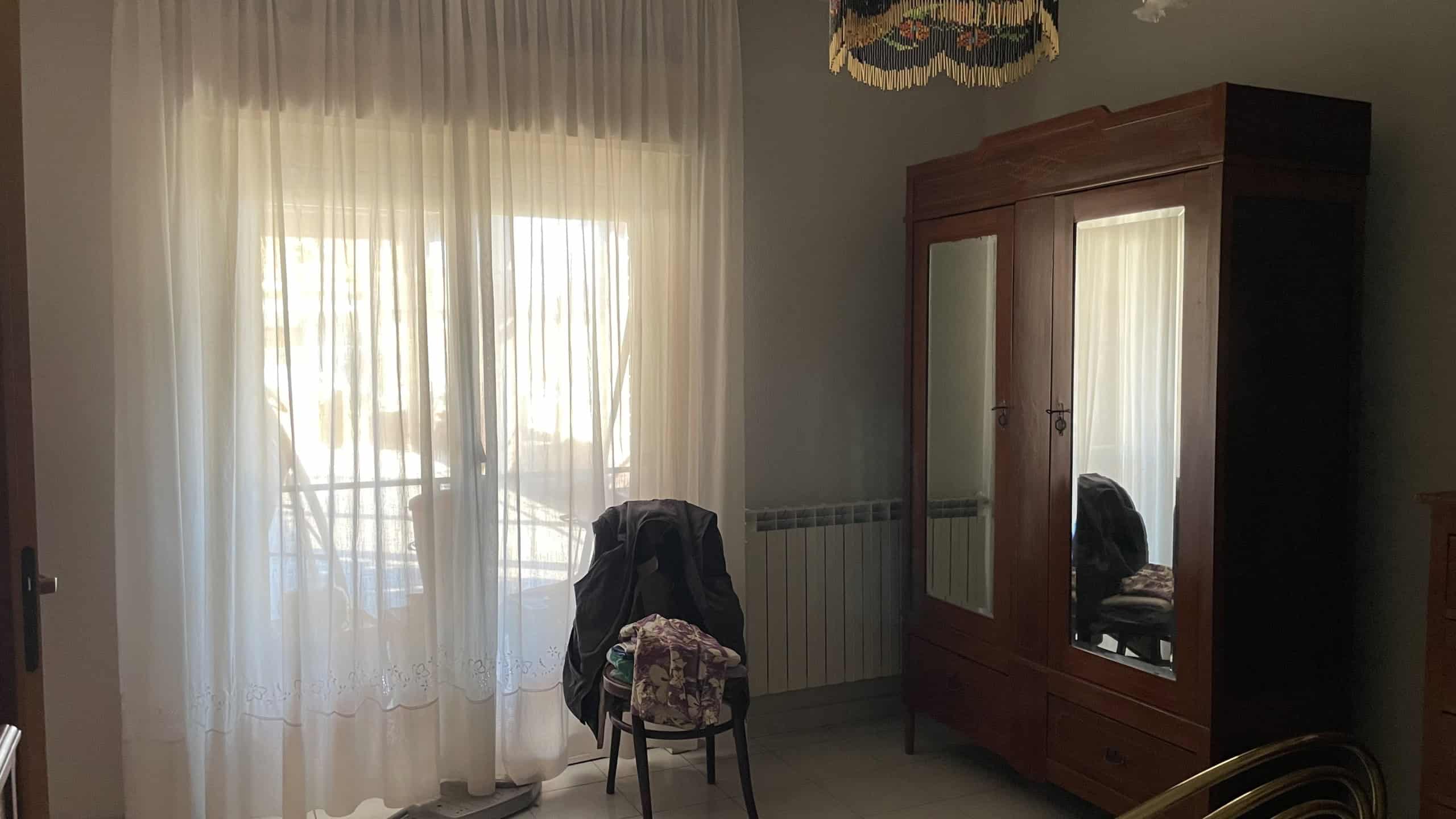 Ampia finestra con tende leggere che lascia entrare molta luce naturale in una stanza arredata con armadio in legno e sedia con copertura floreale, creando un ambiente accogliente e luminoso.