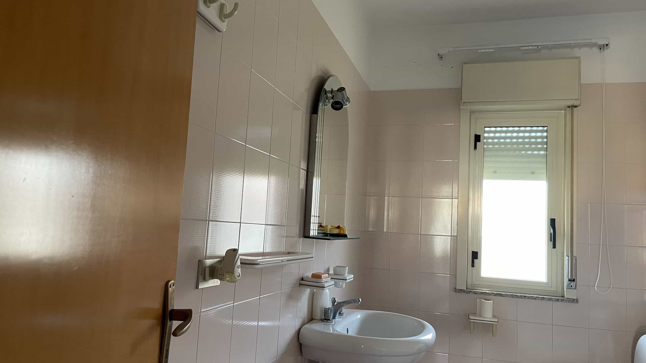 Ampia finestra in bagno con vista luminosa, lavabo bianco e specchio, pareti rivestite con piastrelle beige, ambiente pulito e ben illuminato, ideale per comfort quotidiano.