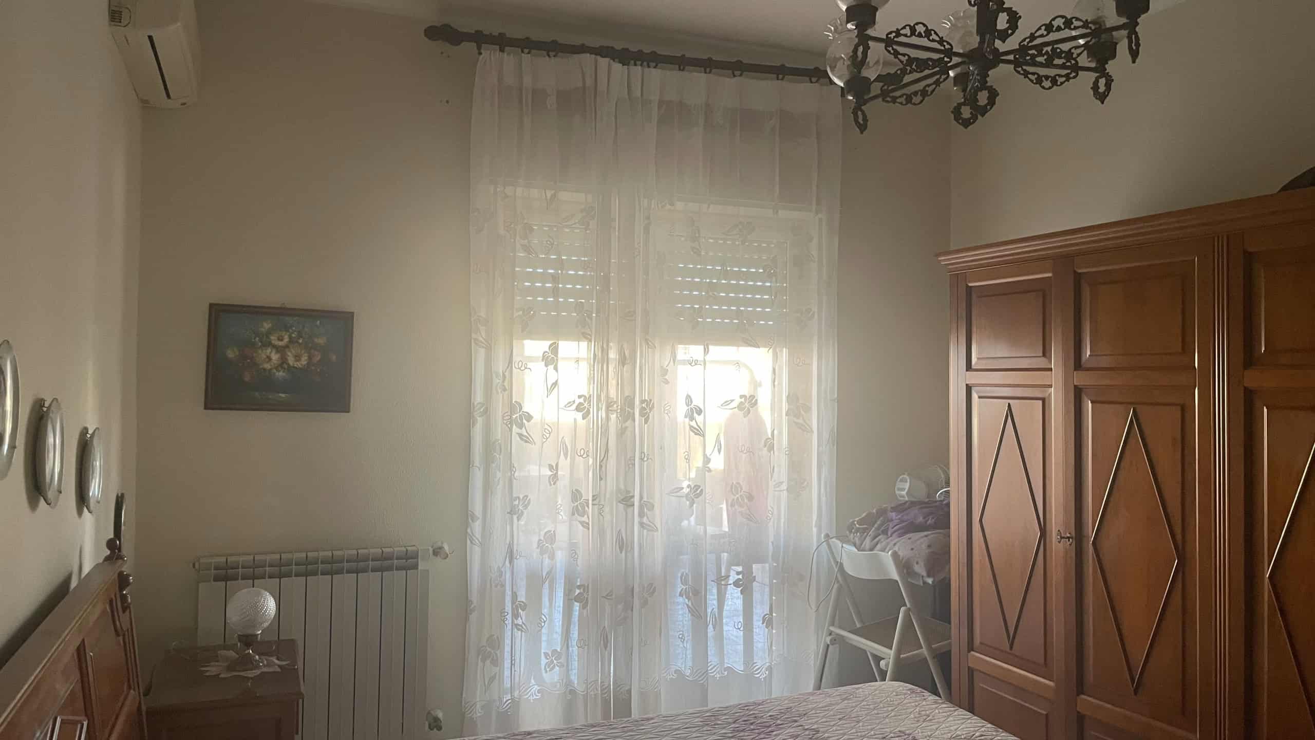 Elegante soggiorno con grande finestra, tende leggere e arredi in legno, tra cui un armadio e un tavolo con sedie, illuminato dalla luce naturale e decorato con un quadro floreale.