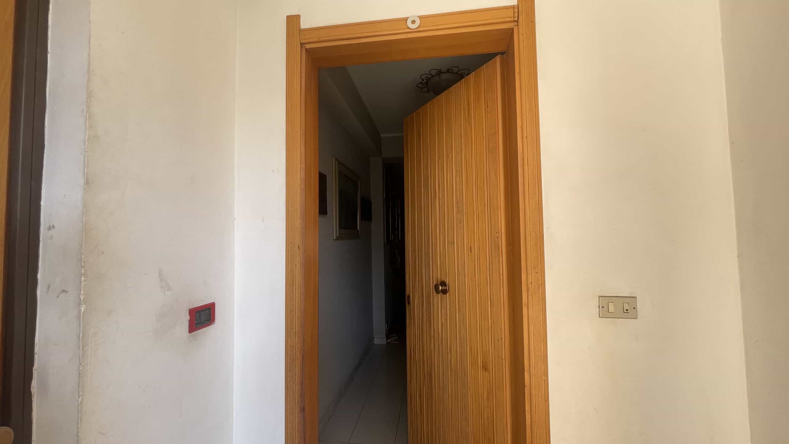 Porta in legno aperta in un ingresso di casa, con pareti bianche e interruttori elettrici visibili, ambiente luminoso e accogliente.
