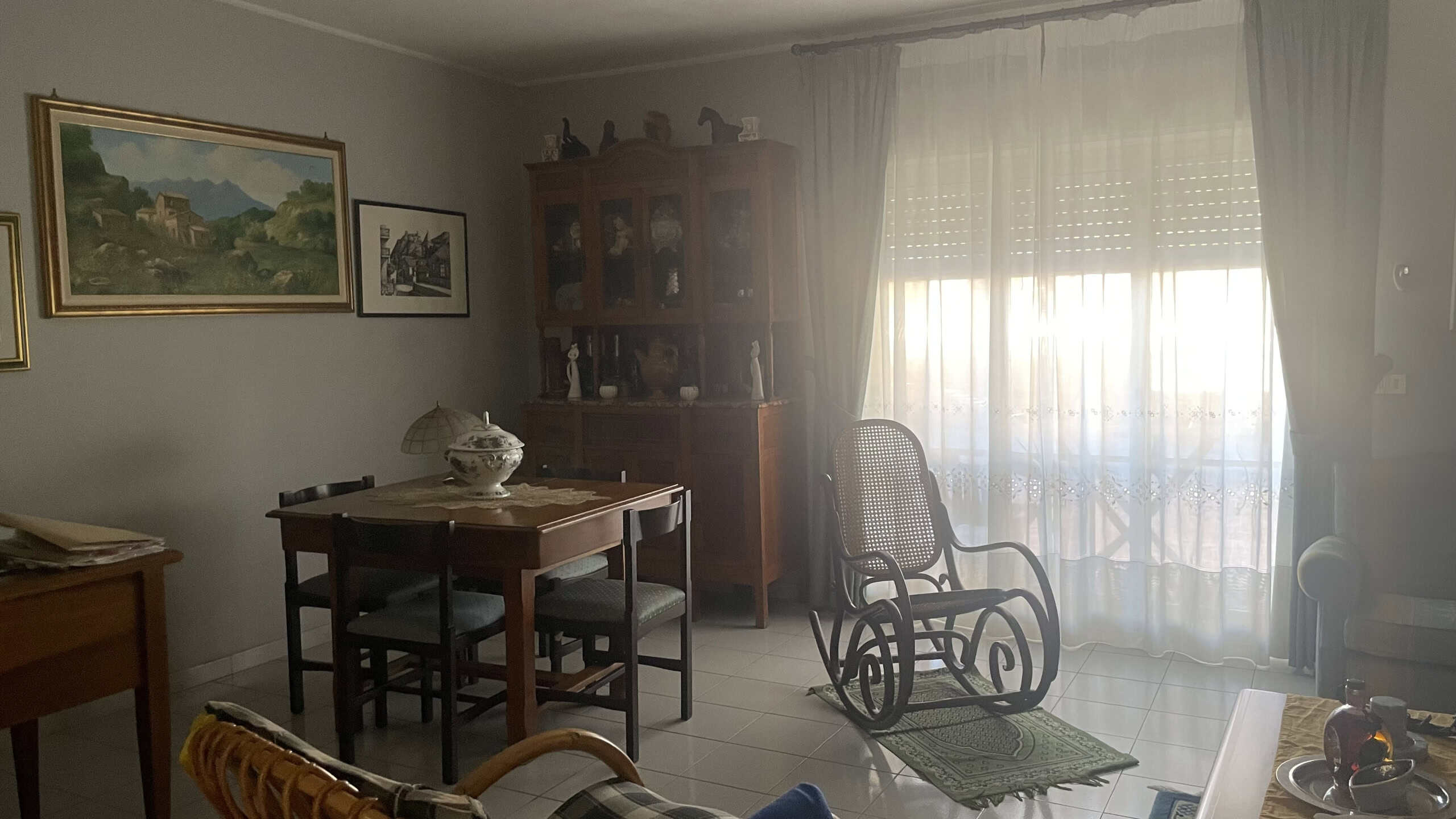 Ampia stanza luminosa con mobili d'epoca, tavolo in legno, sedie, credenza e poltrona, illuminata da luce naturale attraverso grandi finestre con tende leggere, ambiente accogliente e arredato in stil.