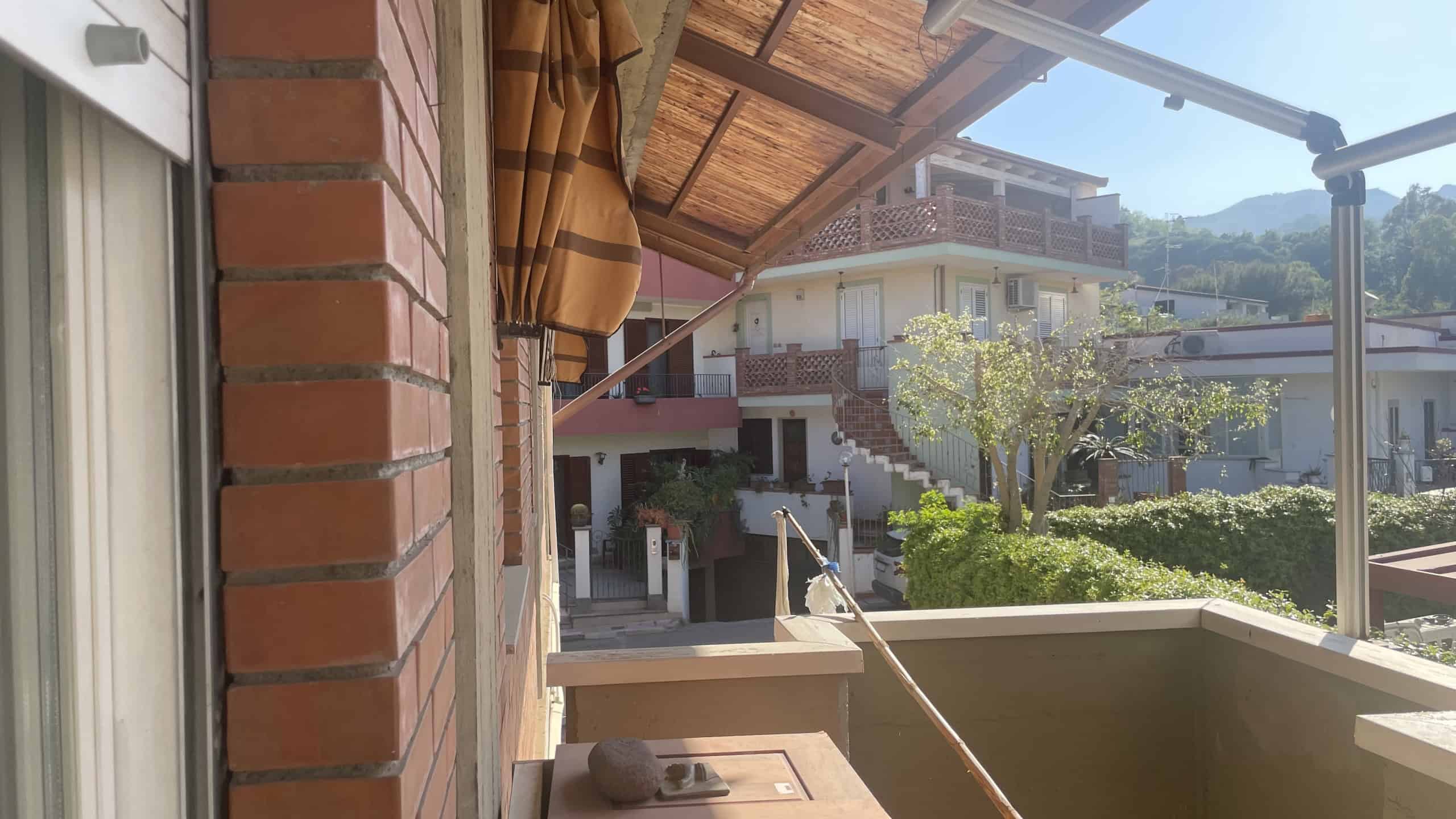 Ampia terrazza con vista su case residenziali e montagne, coperta da tettoia in legno e ombrellone, ideale per relax e convivialità all'aperto.