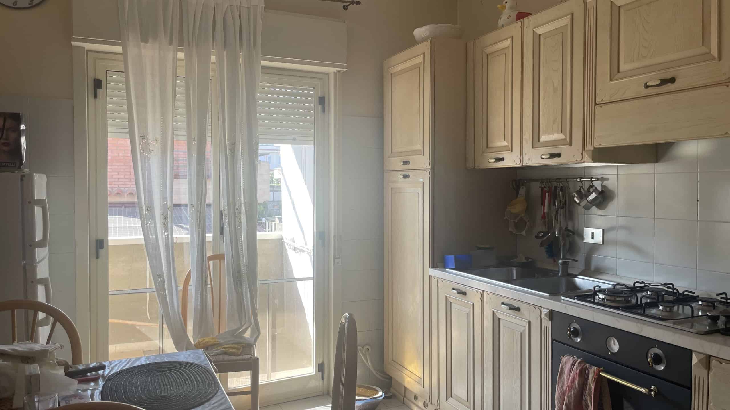 Ampia cucina moderna con armadi in legno chiaro, piano cottura a gas e accessori, illuminata da luce naturale attraverso una porta finestra con tende leggere, ideale per vivere in comfort.