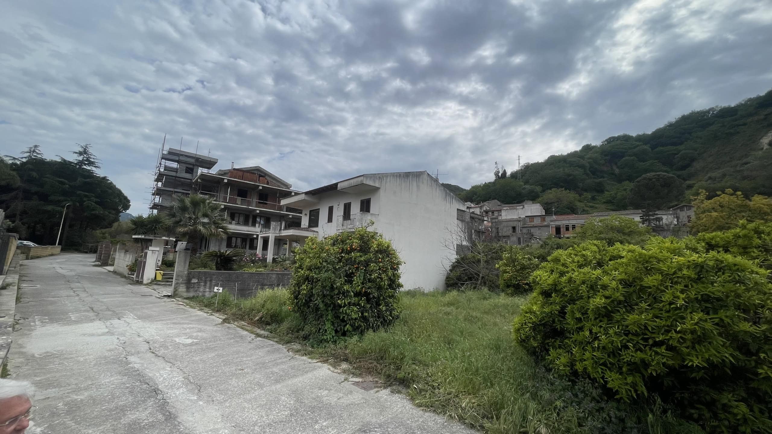 Edificio residenziale in costruzione in zona collinare con cielo nuvoloso, piante verdi e strada asfaltata, immagine di un'area in sviluppo immobiliare a NextCasa.