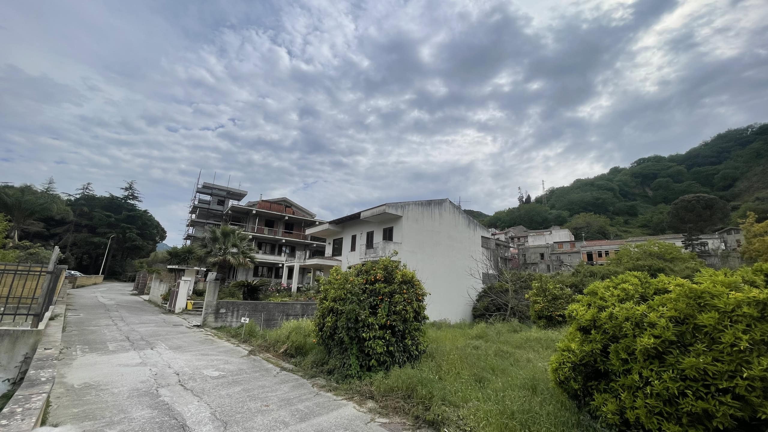 Ampia strada in salita con case residenziali moderne e tradizionali, circondata da vegetazione verde e colline sullo sfondo, sotto un cielo nuvoloso, in un quartiere tranquillo e panoramico.